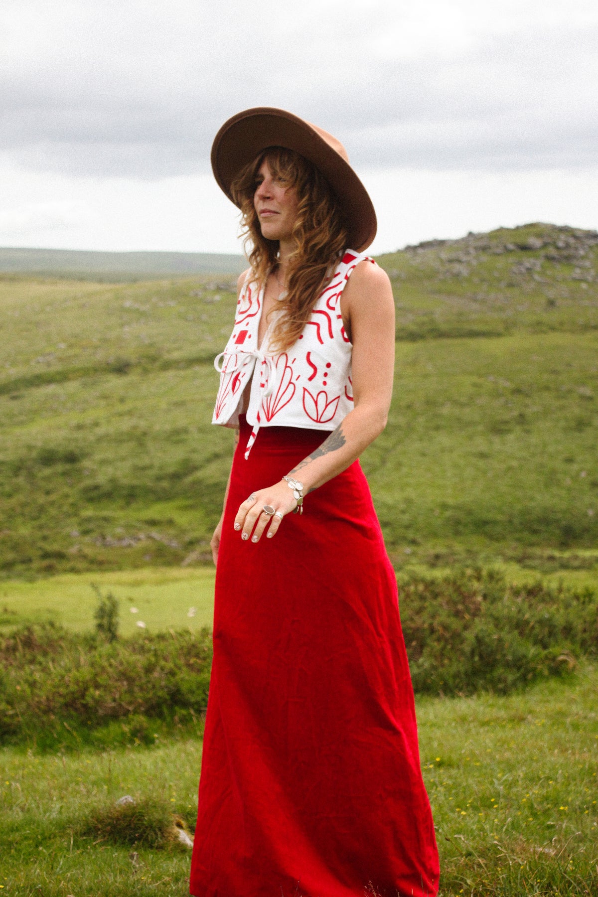 A woman standing outdoors in a grassy landscape wearing a wide-brimmed brown hat, a white sleeveless top with red abstract line patterns, and a long red skirt.
