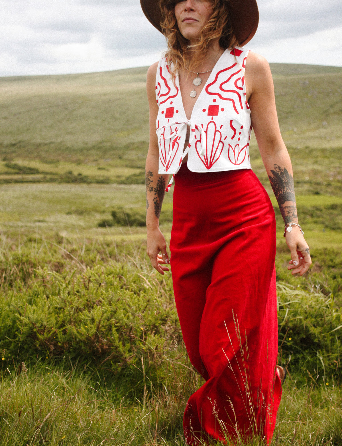 A woman wearing a sleeveless white vest with red abstract patterns and a matching red maxi skirt, walking outdoors in a grassy field under an overcast sky.