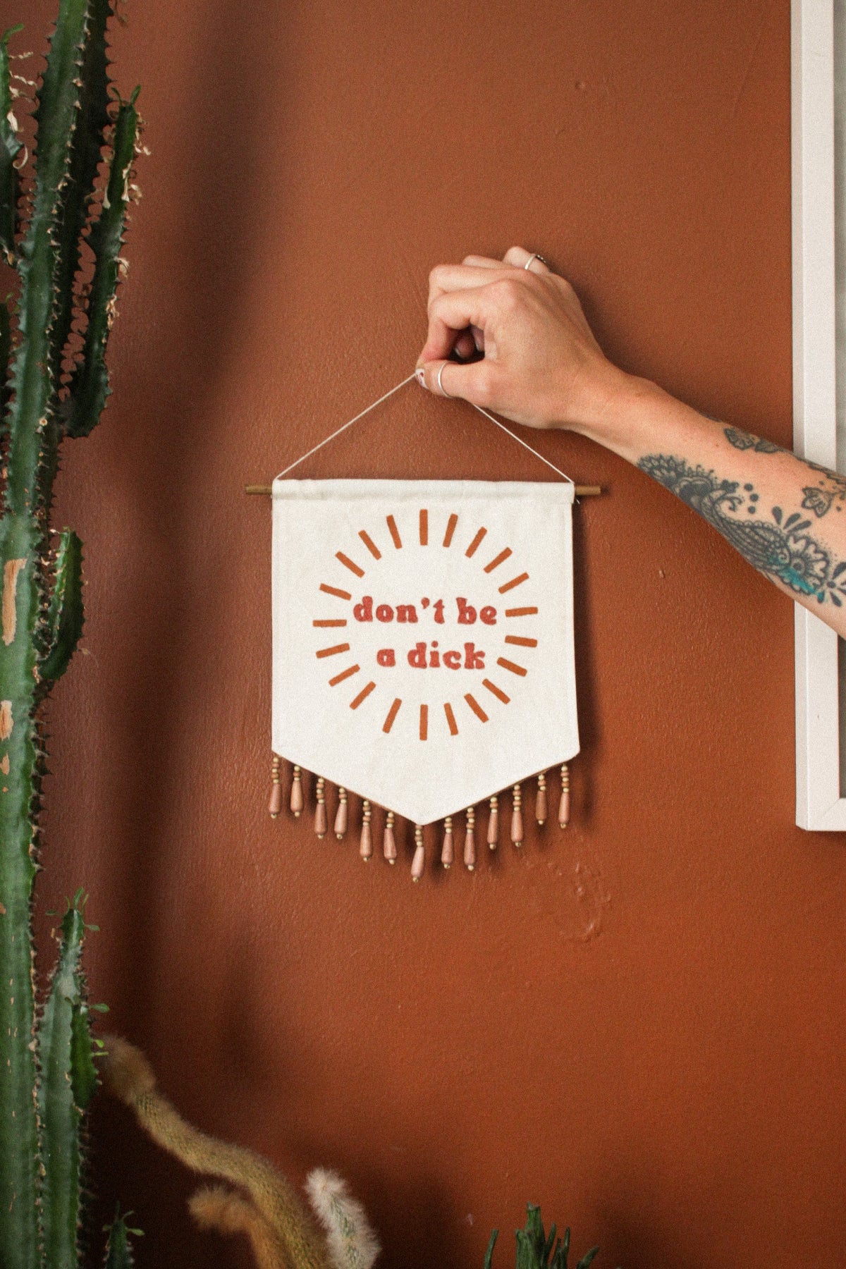 A hand with a tattoo holds a small white fabric pennant banner with orange text reading 'don't be a dick' and orange radiating lines, against a terracotta wall with a cactus on the left.
