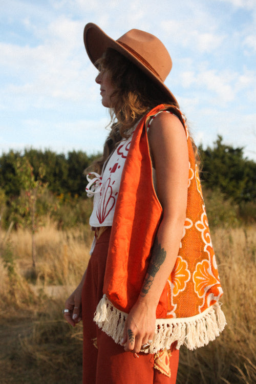 A woman in a bohemian-style outfit stands in an outdoor field. She is wearing a wide-brimmed brown hat, a white top with red hand painted floral patterns, rust-orange trousers, and a bright orange boho style bag with cream fringe and floral embroidery.