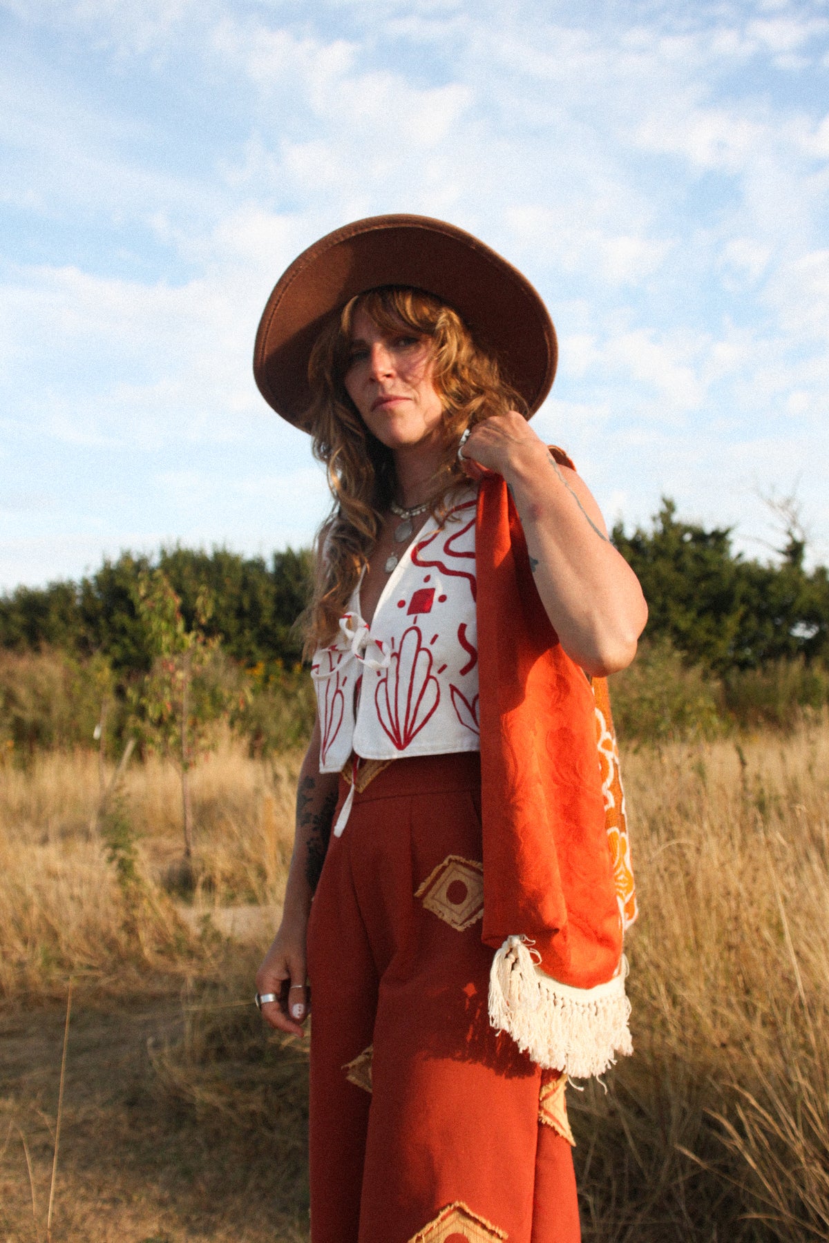 A woman stands outdoors in a grassy field under a blue sky, wearing a wide-brimmed brown hat, a white top with red abstract patterns, rust-orange pants, and a bright orange fringed wrap draped over her shoulder.