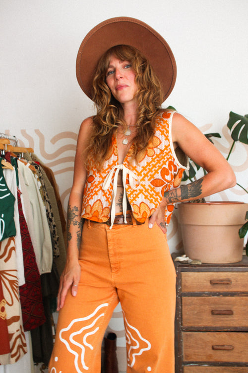 A woman wearing a waistcoat in orange and white boho patterns with drawstring ties, matching orange pants with white western-style line drawings, and a brown wide-brim hat.