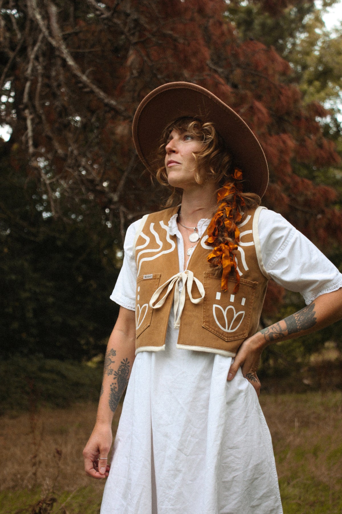 A person outdoors wearing a wide-brim brown hat, a white dress, and a tan hand painted vest with white bohemian style patterns and a off white ties