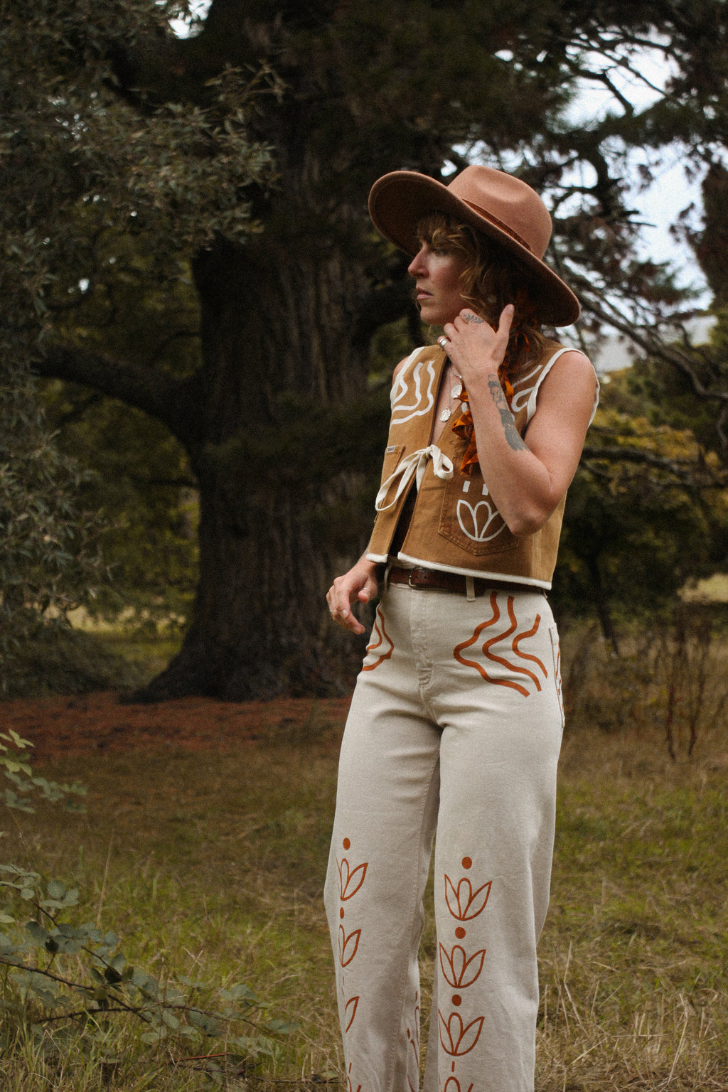 A person standing outdoors in a grassy wooded area wearing a tan wide-brim hat, a sleeveless tan vest with white and rust-orange abstract bohemian painting, cream wide-leg pants with rust-orange embroidered leaf motifs, and a dark belt.