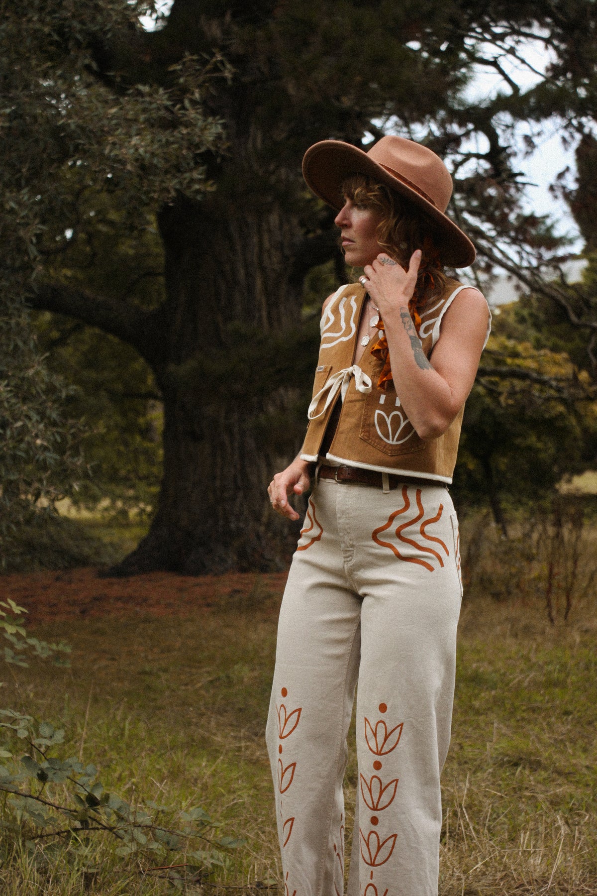A person standing outdoors in a grassy wooded area wearing a tan wide-brim hat, a sleeveless tan vest with white and rust-orange abstract bohemian painting, cream wide-leg pants with rust-orange embroidered leaf motifs, and a dark belt.