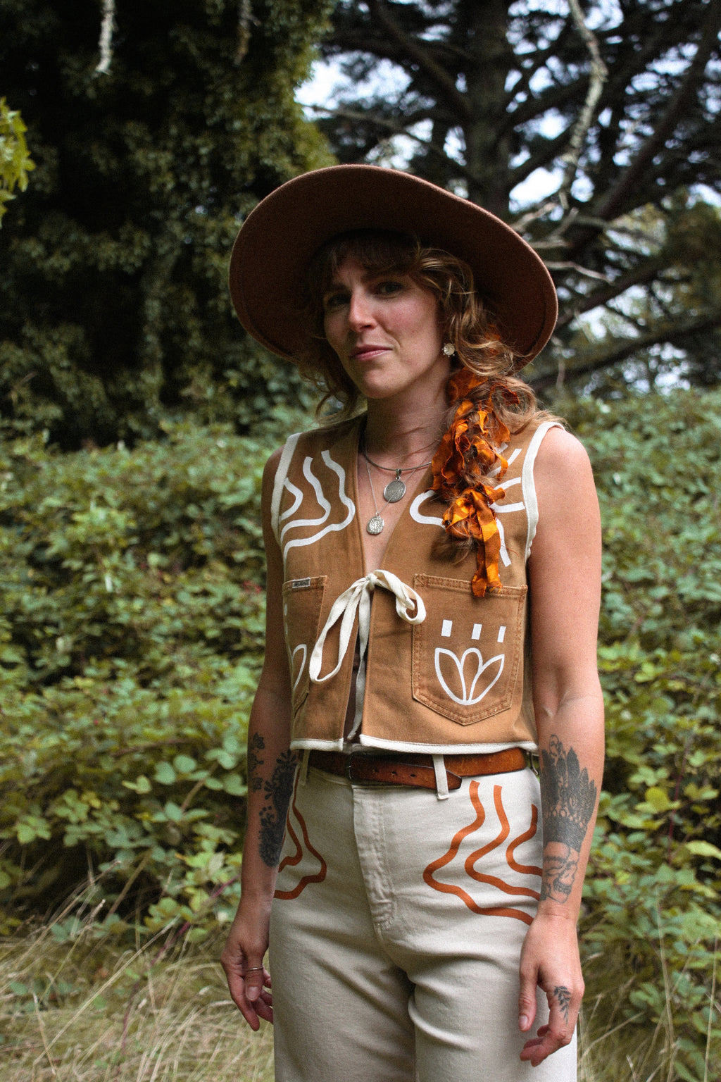 A woman wearing a brown wide-brim hat, a tan sleeveless vest with white and orange hand-drawn style patterns, and beige pants, standing outdoors in front of green trees and bushes.