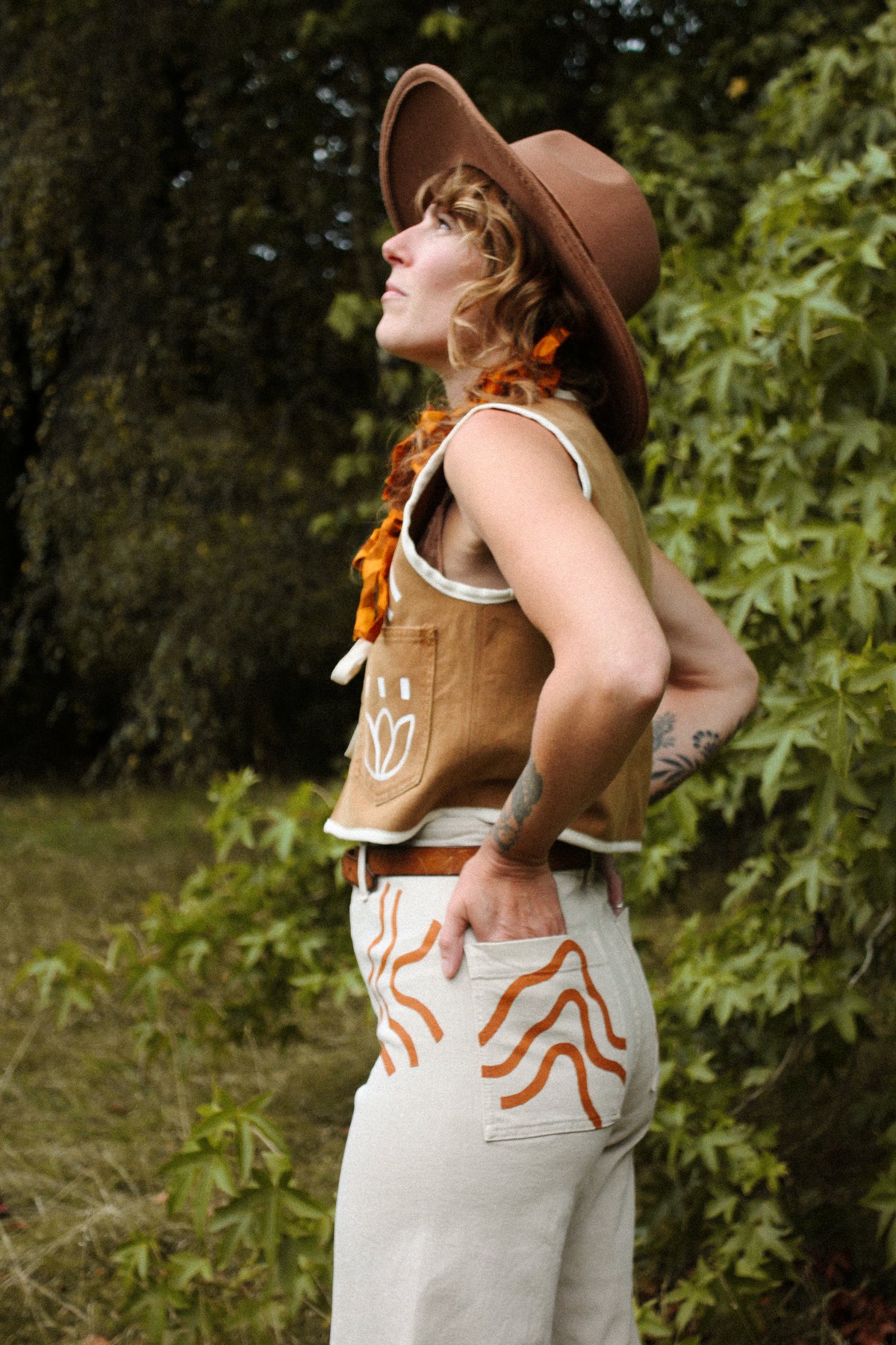 A person wearing a brown wide-brim hat, a brown sleeveless vest with white geometric embroidery, an orange patterned scarf, and light beige pants with orange zigzag embroidery, standing outdoors in front of green foliage.