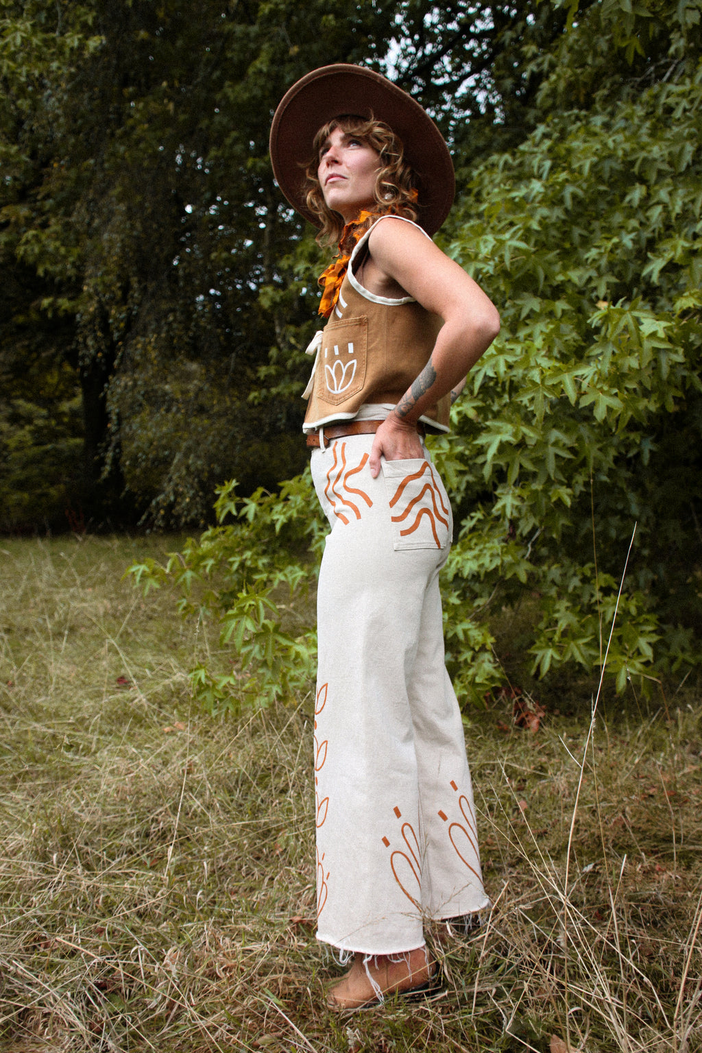 A woman stands outdoors in a grassy wooded area wearing a bohemian-style outfit consisting of a brown wide-brim hat, a brown vest with white hand-drawn style graphics, a bright orange scarf, and off-white wide-leg pants with orange tribal-style patterns.