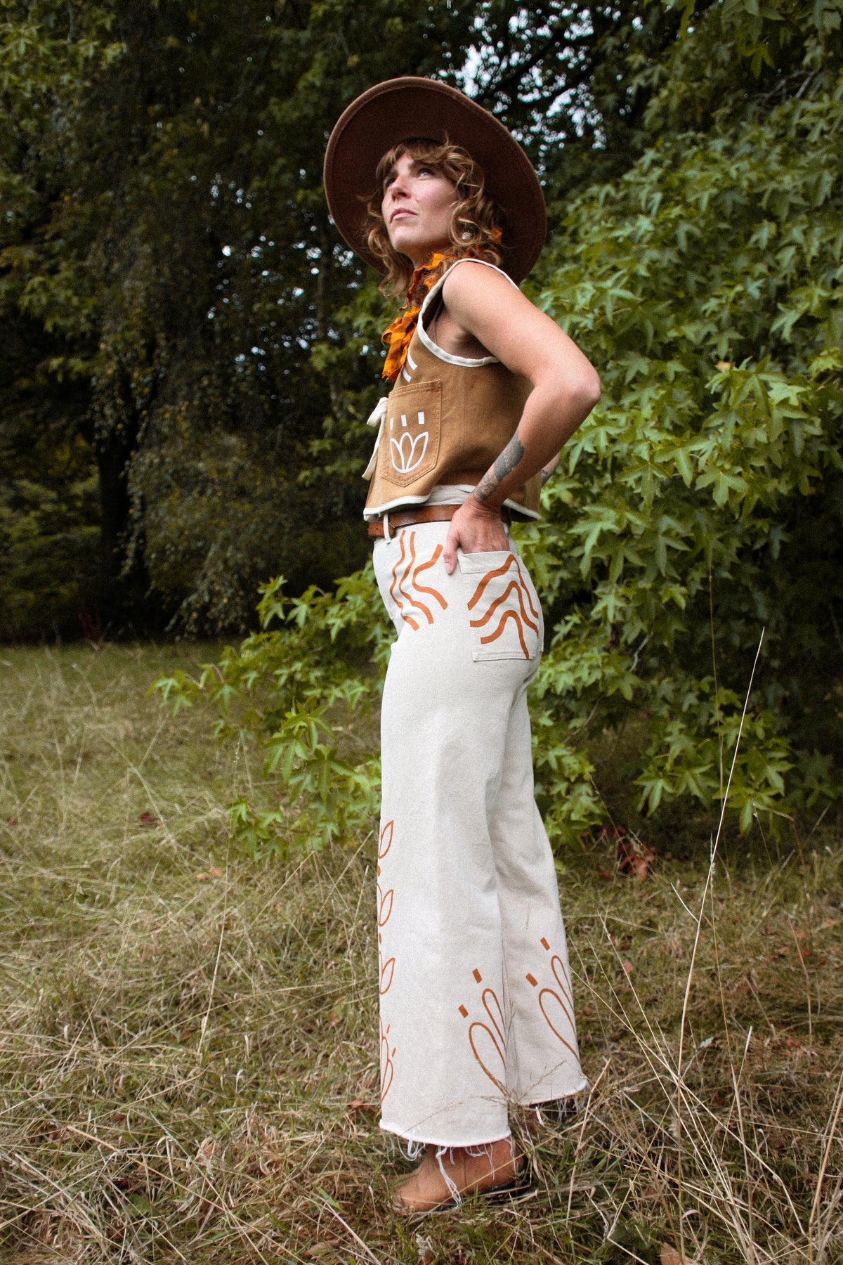 A woman stands outdoors in a grassy wooded area wearing a bohemian-style outfit consisting of a brown wide-brim hat, a brown vest with white hand-drawn style graphics, a bright orange scarf, and off-white wide-leg pants with orange tribal-style patterns.