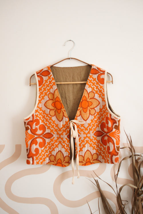 An orange and beige patterned vest made from a 70's curtain with a tie-front closure hanging on a wooden hanger against a light beige wall.