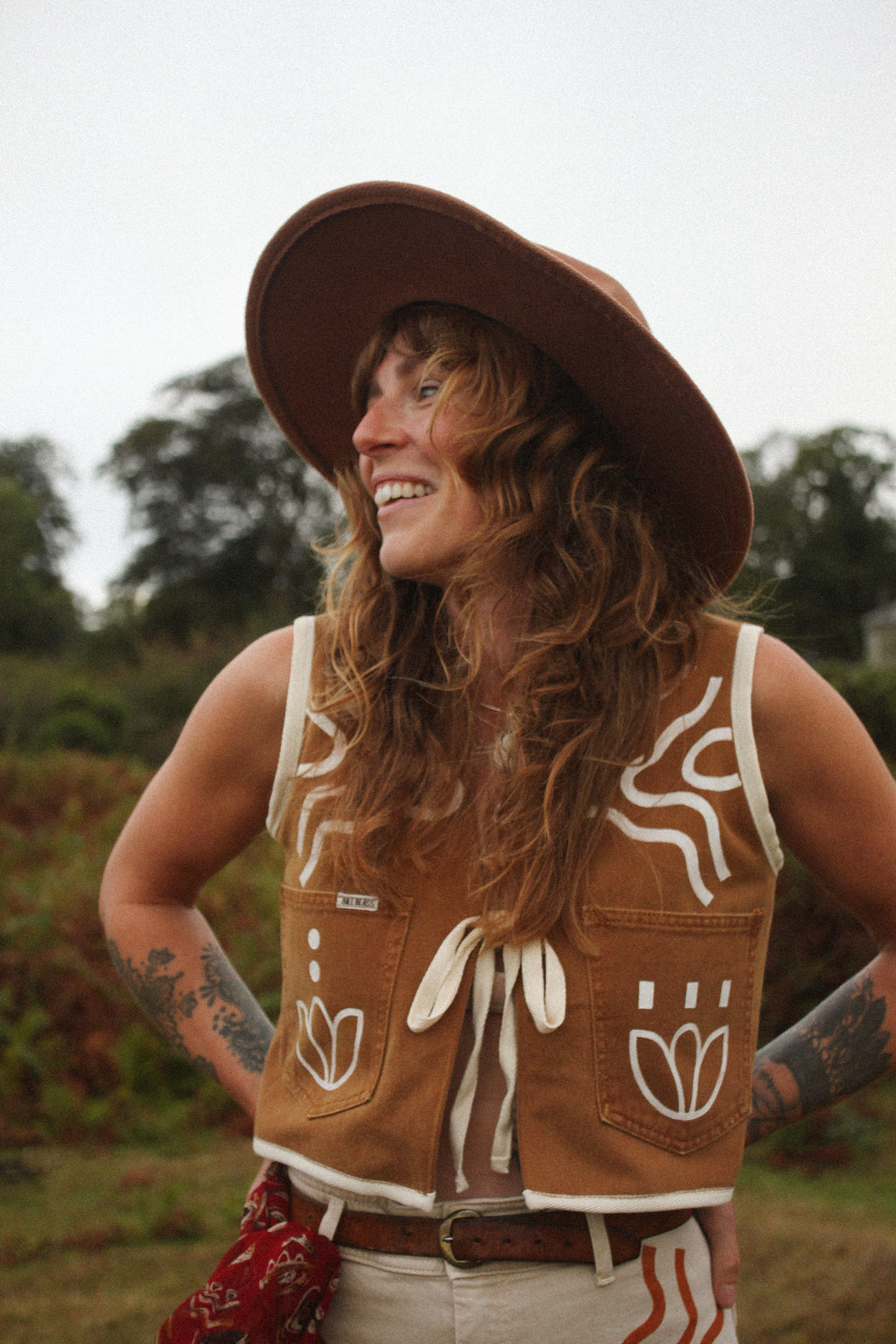 A woman outdoors wearing a brown wide-brim hat and a brown sleeveless vest with white decorative graphics and a front tie, paired with light-colored pants. She is smiling and looking to the side against a blurred natural background.