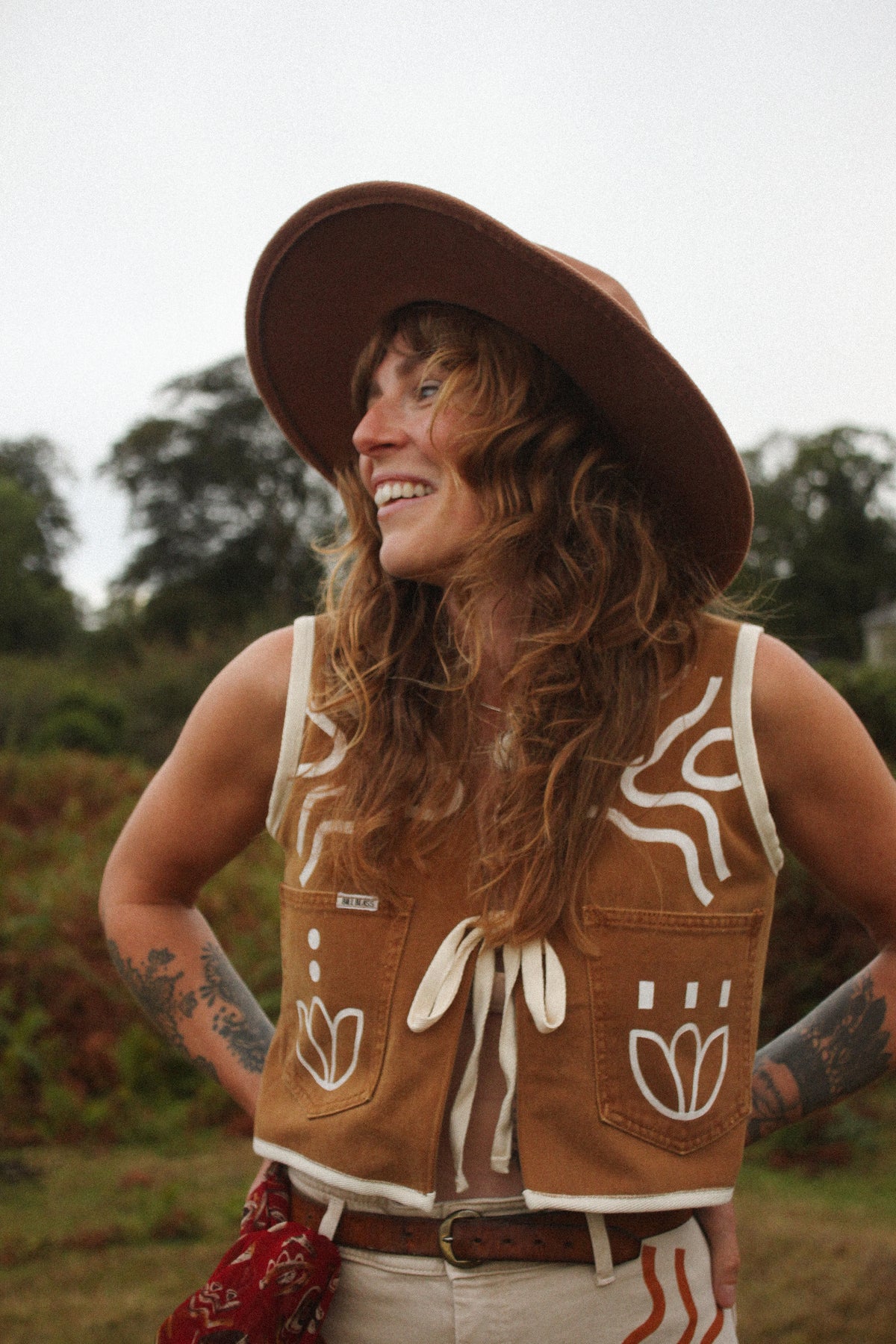 A woman outdoors wearing a brown wide-brim hat and a brown sleeveless vest with white decorative graphics and a front tie, paired with light-colored pants. She is smiling and looking to the side against a blurred natural background.