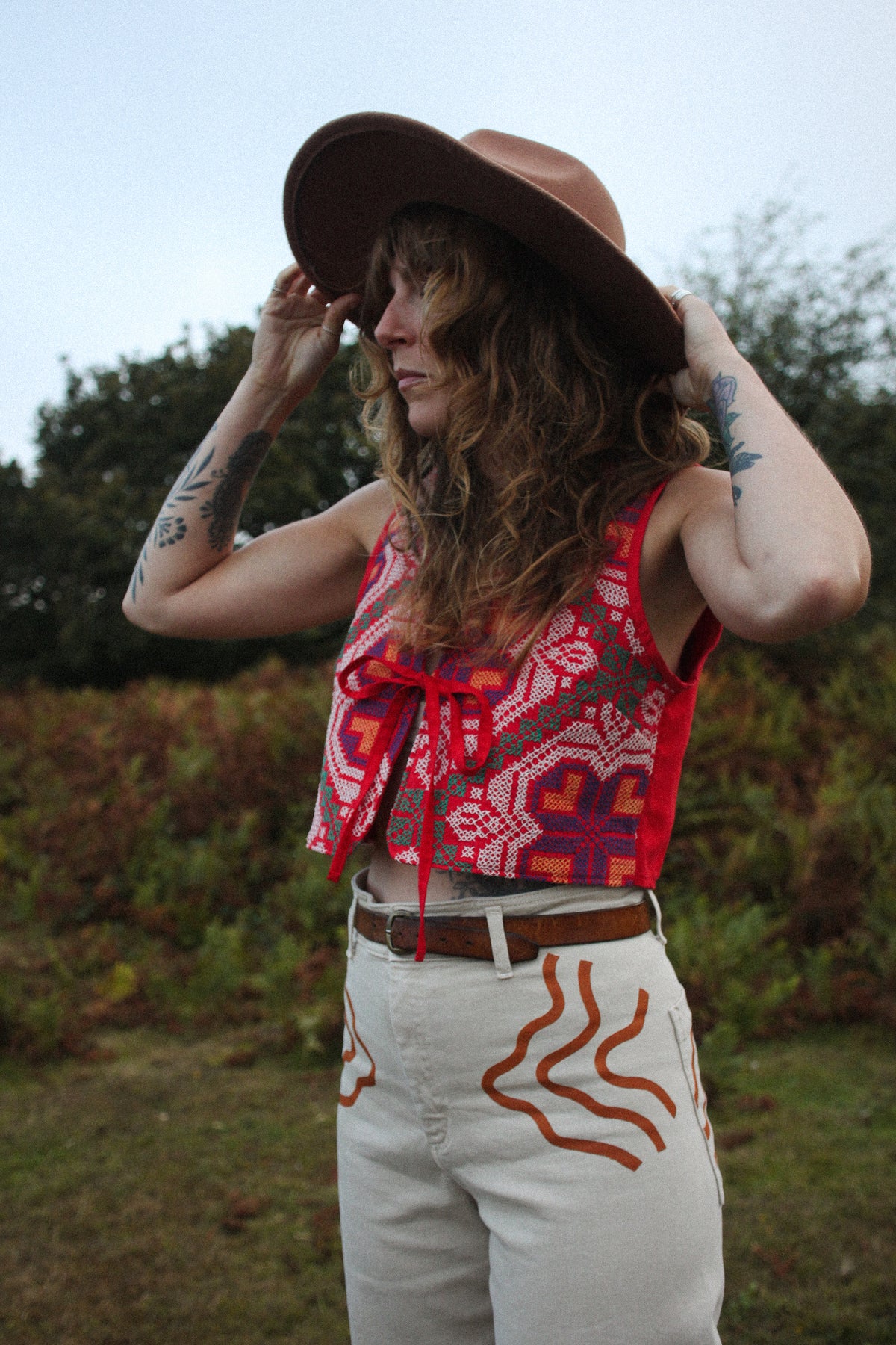 A woman outdoors wearing a brown wide-brim hat, a sleeveless red and purple geometric patterned crop top with red side ties, and off-white pants with orange abstract line details.