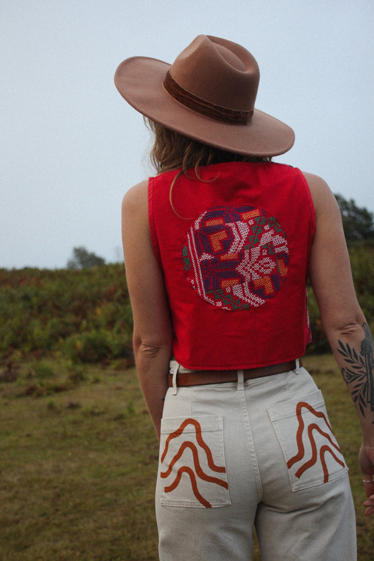 A person standing outdoors in a grassy field, viewed from behind, wearing a brown wide-brim hat, a bright red sleeveless top with a large multicolor geometric circular print on the back, and off-white pants with orange painted patterns on the back pockets.