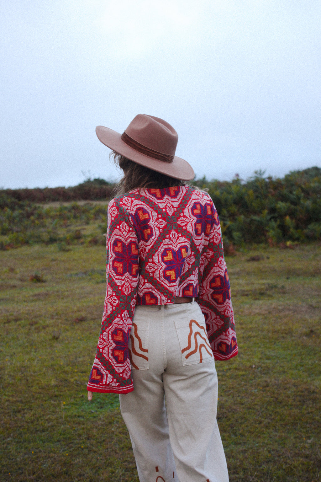 A person standing outdoors in a grassy field, viewed from behind, wearing a brown wide-brim hat, a red bell sleeve 70s patterned long-sleeve cropped top, and cream-colored pants.