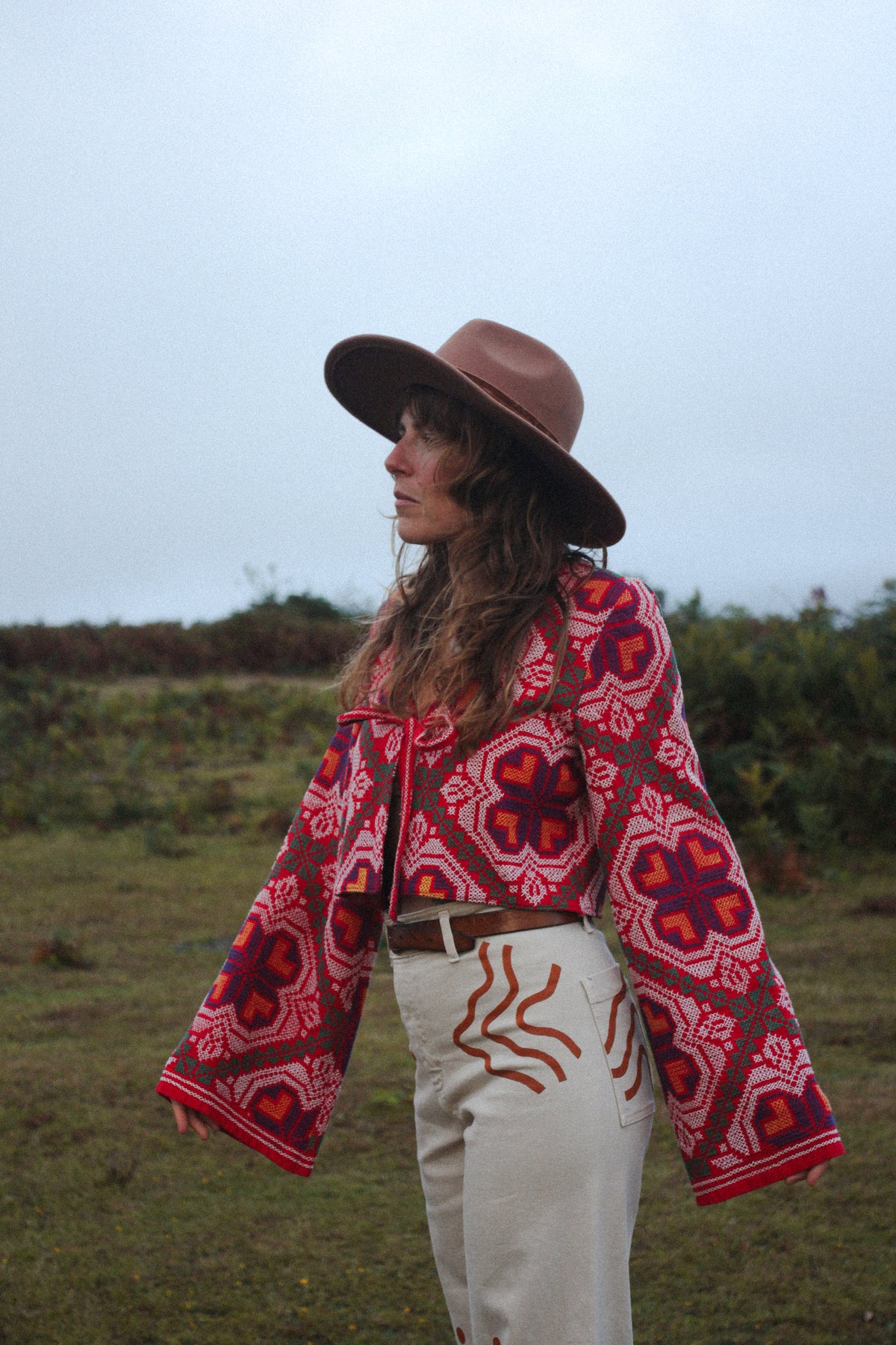 A woman wearing a brown wide-brim hat, a red and black patterned cropped jacket, and cream pants stands outdoors in a grassy field under an overcast sky.