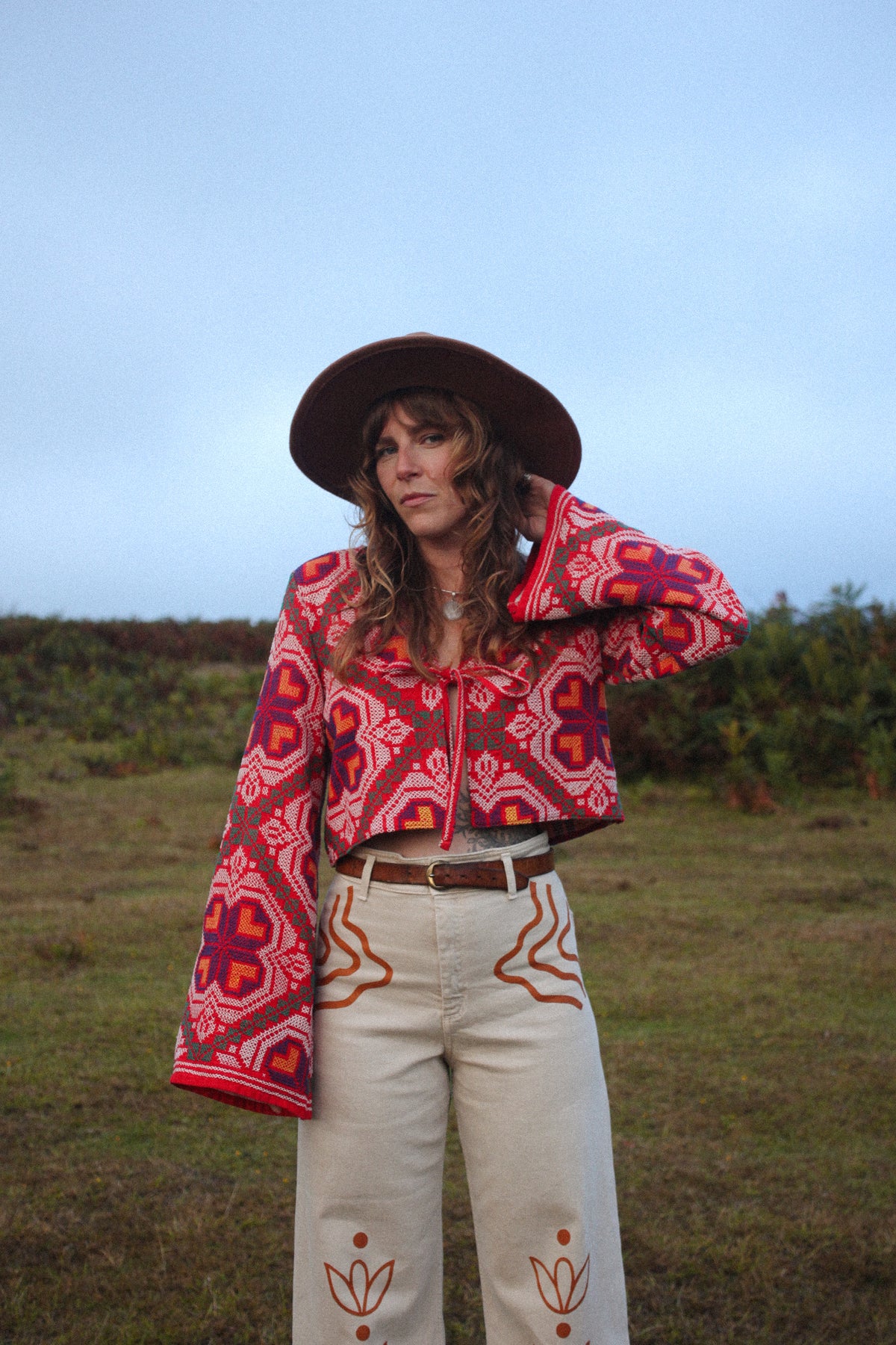 A woman wearing a bohemian-style outfit stands outdoors in a grassy field under a pale blue sky. She is wearing a wide-brimmed brown hat, a red, 70s patterned cropped jacket, and cream-colored pants with orange painted patterns.