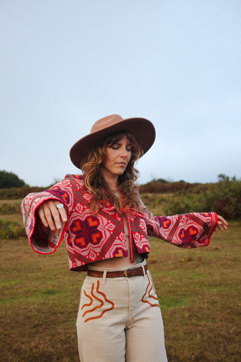 A woman wearing a bohemian-style patterned crop top, wide-leg beige pants, and a brown wide-brim hat, standing in an outdoor grassy field.