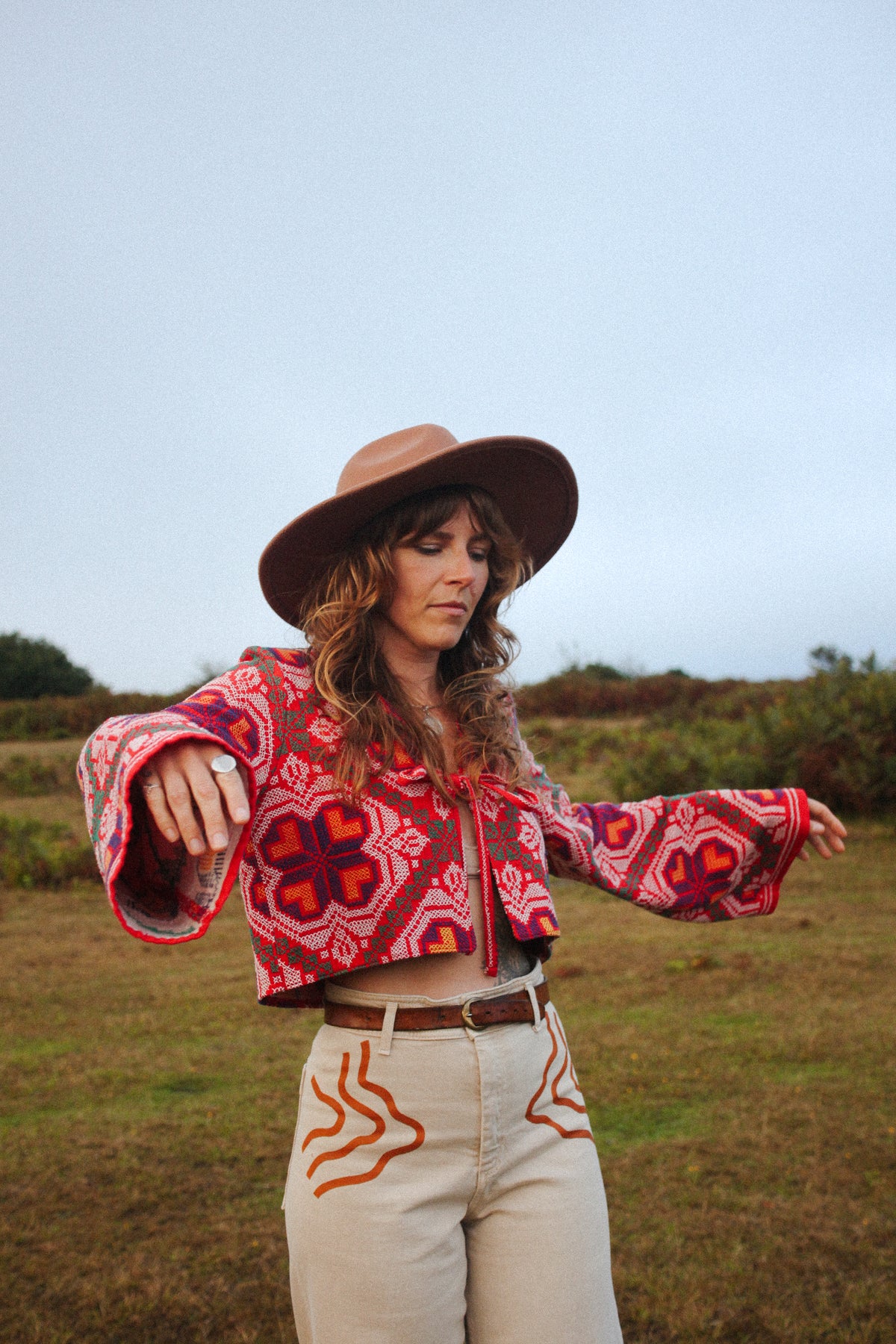 A woman wearing a bohemian-style patterned crop top, wide-leg beige pants, and a brown wide-brim hat, standing in an outdoor grassy field.