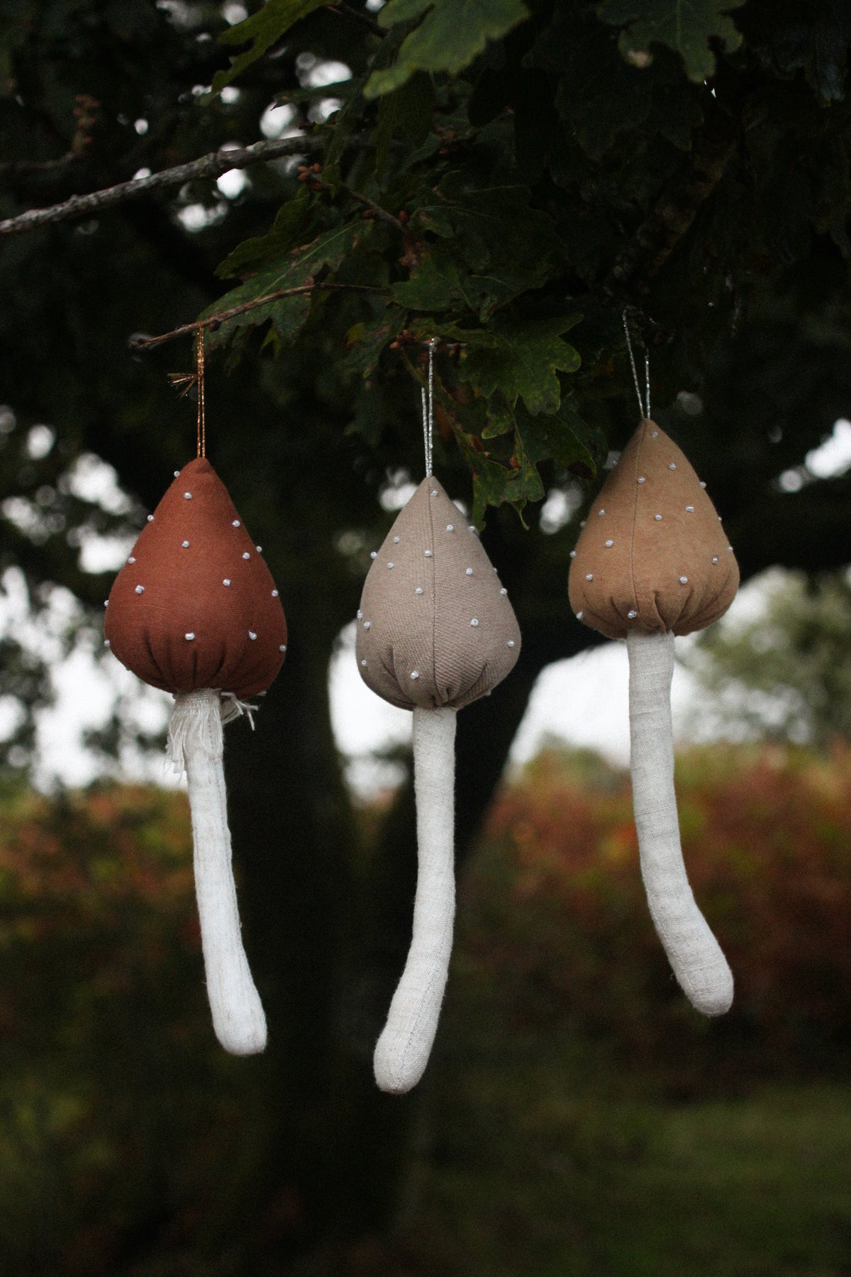 Three mushroom-shaped hanging ornaments made of linen and yarn are suspended from tree branches outdoors. The ornaments have brown, taupe, and beige caps with white yarn stems and small white dots, set against a blurred background of green leaves and foliage.