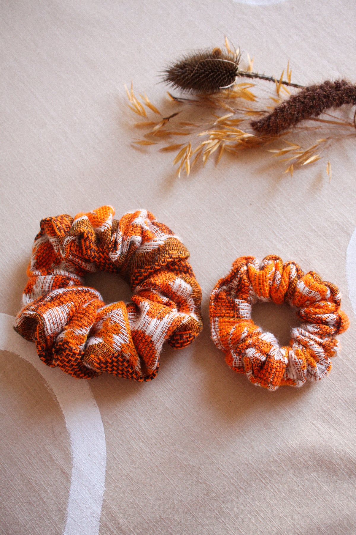 Two fabric hair scrunchies in orange, brown, and white plaid patterns, displayed on a beige fabric surface with dried botanical props.