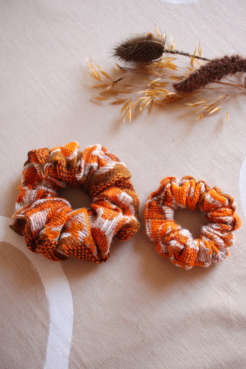 Two fabric hair scrunchies in orange, brown, and white plaid patterns, displayed on a beige fabric surface with dried botanical props.