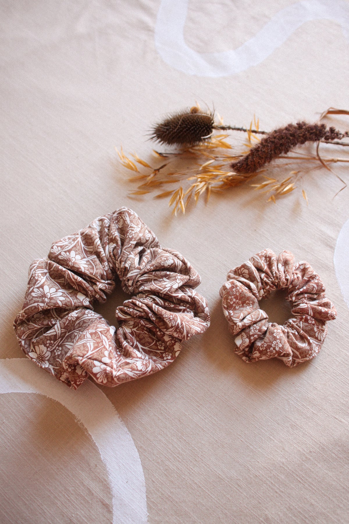 Two dusty rose velvet scrunchies with a white marble pattern, displayed on a beige fabric background with dried decorative grasses.