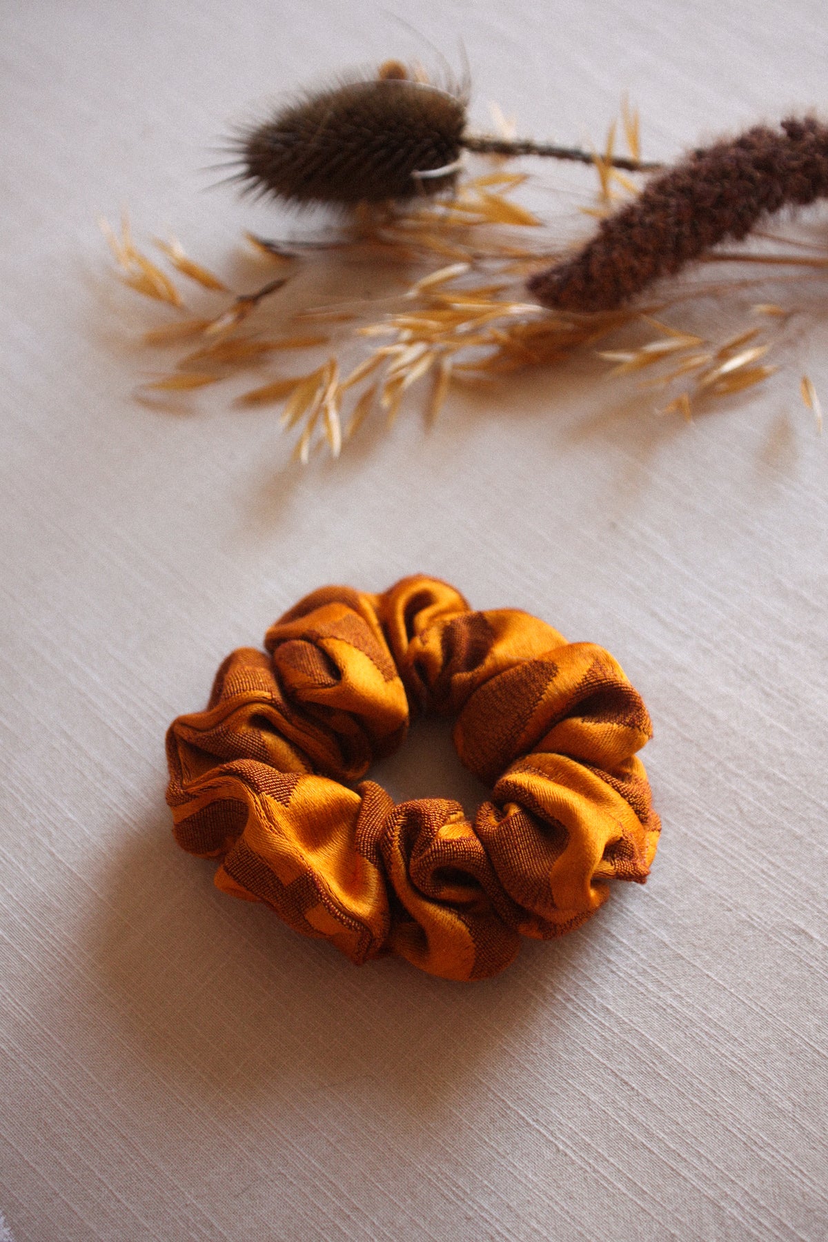 An orange satin scrunchie with a ruffled texture, displayed on a light beige fabric background with dried decorative grasses and a thistle seed pod in the upper background.