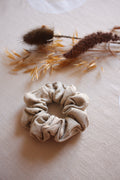 A beige fabric scrunchie made of textured, gathered material, styled on a light beige linen surface with dried botanicals in the background.
