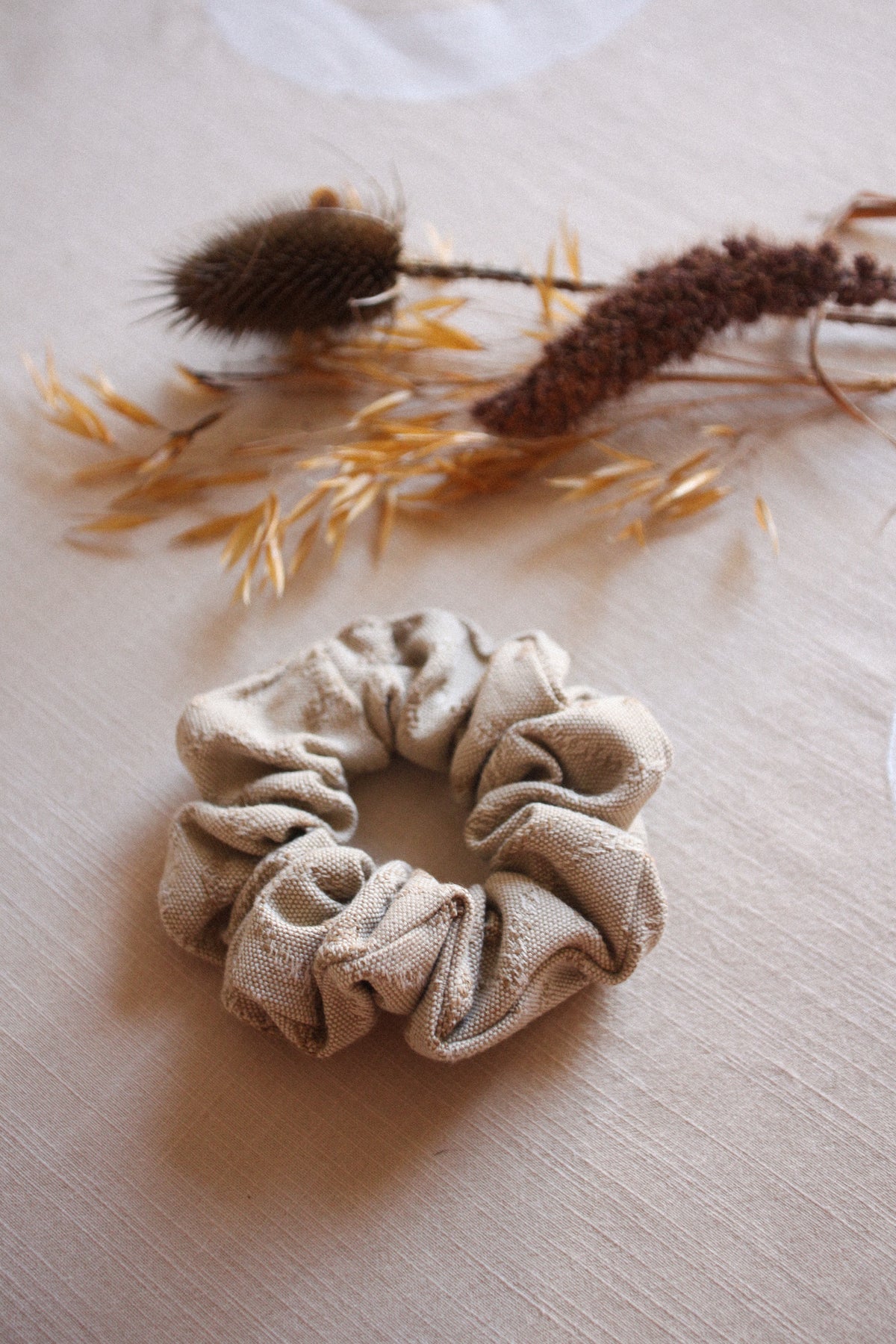 A beige fabric scrunchie made of textured, gathered material, styled on a light beige linen surface with dried botanicals in the background.