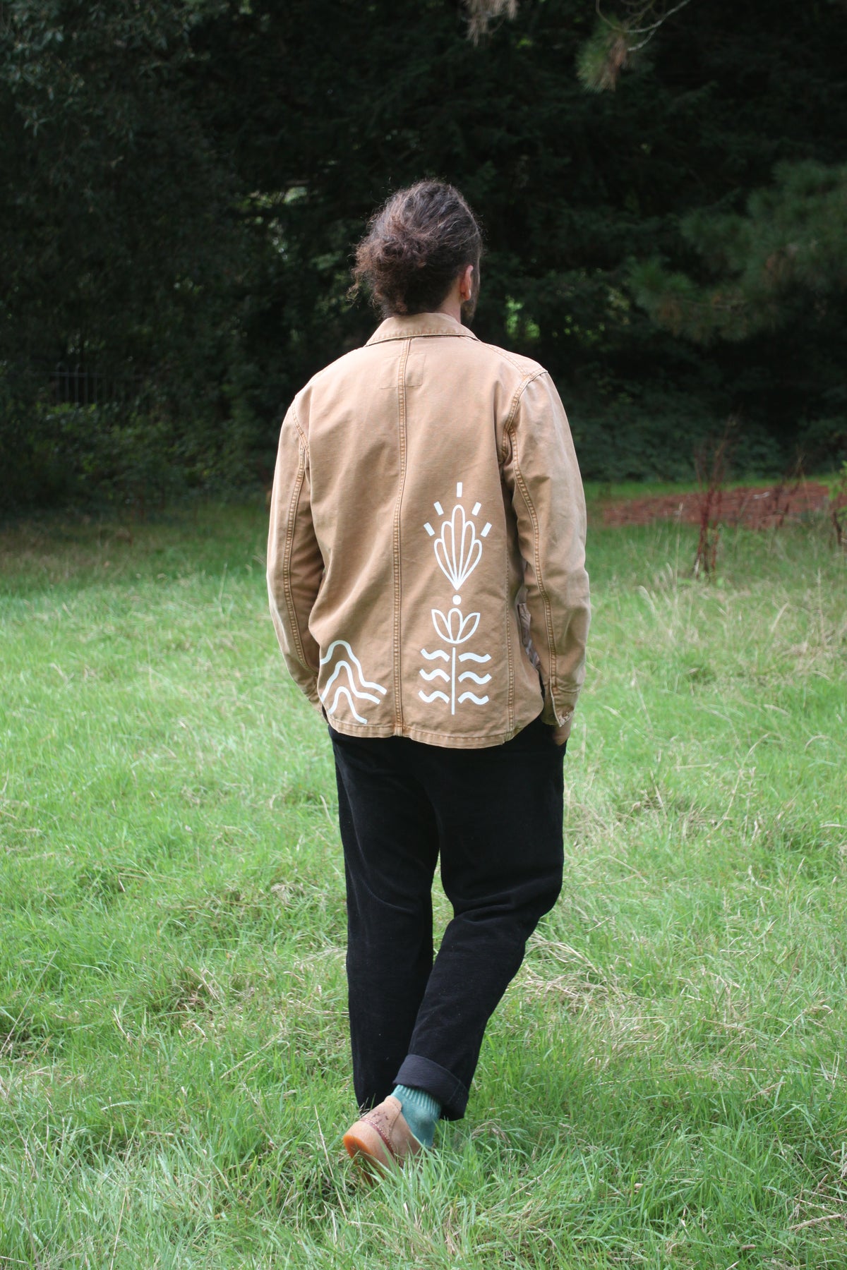 A man viewed from behind walking in a grassy field, wearing a tan utility-style jacket with white hand-painted floral and leaf motifs on the back, paired with black pants and brown shoes.