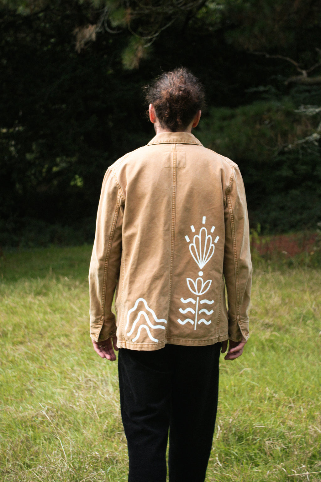A man viewed from behind wearing a tan utility jacket with white hand-drawn style floral and mountain motifs on the back, standing in a grassy outdoor area with trees in the background.
