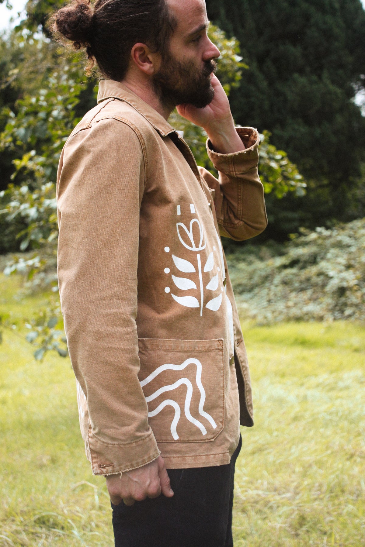 A man wearing a tan utility jacket with white abstract graphic prints on the front, standing outdoors in a grassy field with trees in the background.