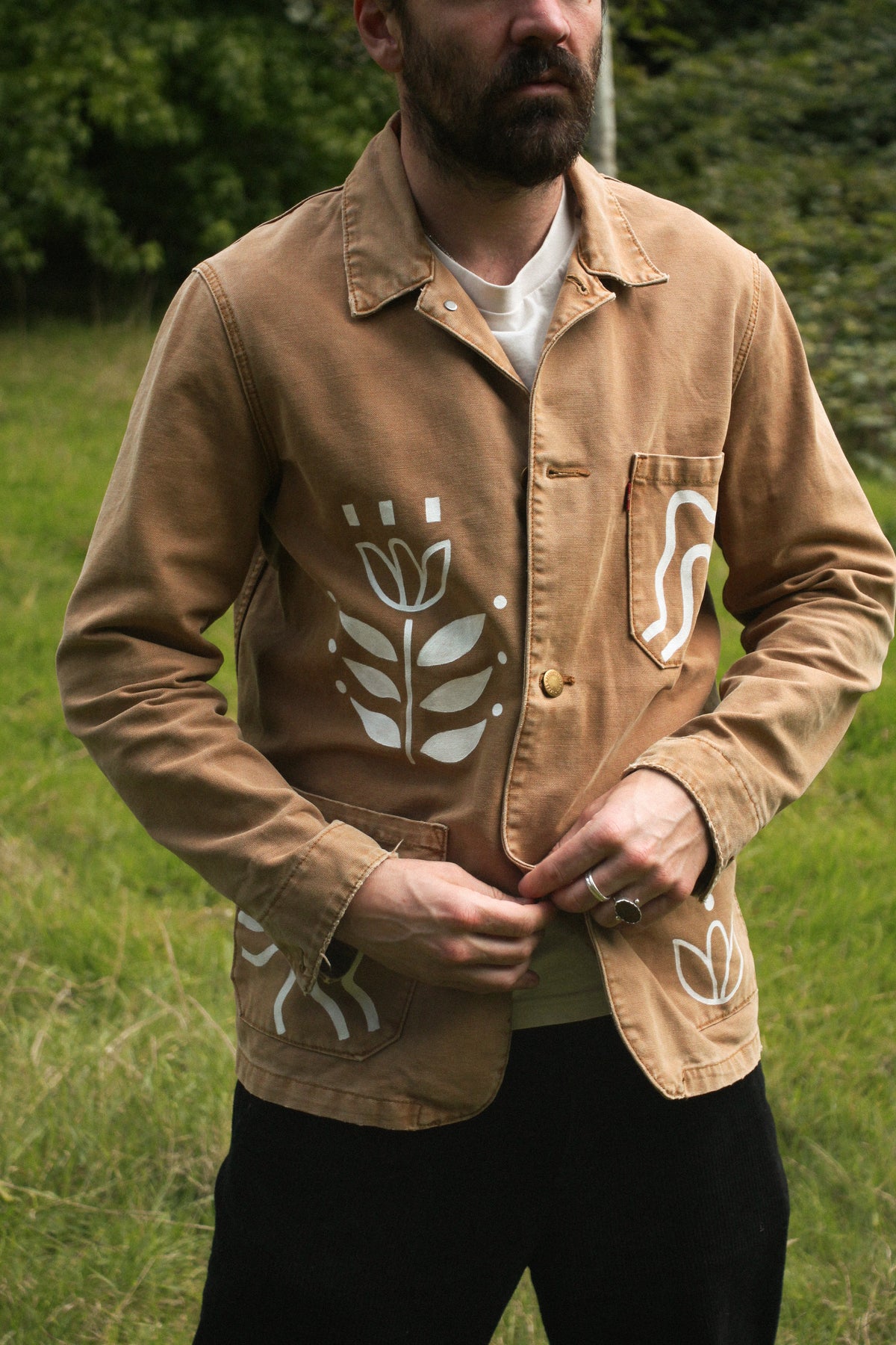 A man wearing a tan utility jacket with white hand-drawn style tulip motifs, standing outdoors in a grassy field.