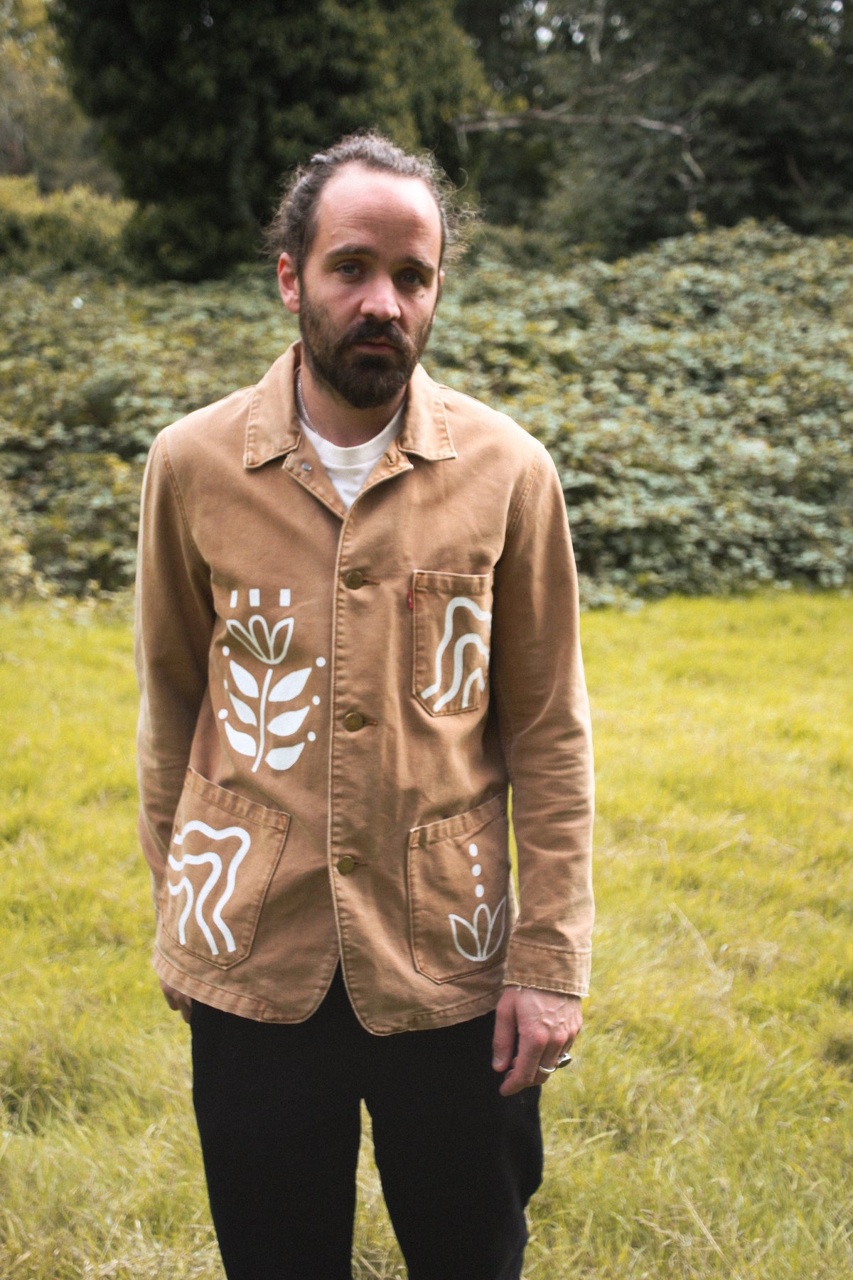 A man standing outdoors in a grassy field wearing a tan utility jacket with white hand-drawn style botanical and abstract patterns on the front pockets, paired with black pants.