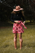 A woman standing in a grassy field wearing a brown wide-brim hat, a black long-sleeve top, a red and pink paisley patterned wrap skirt, and brown cowboy boots.