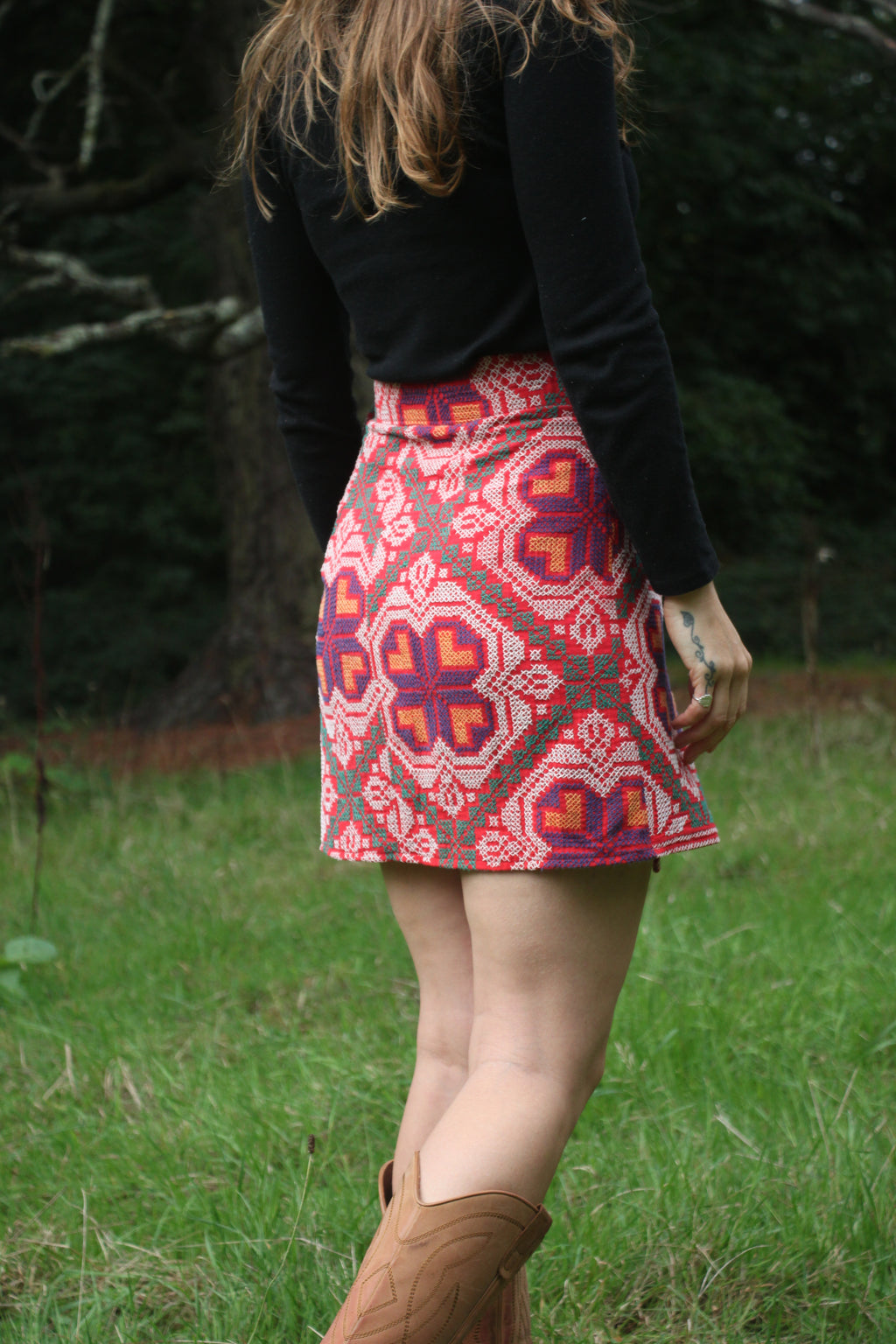 A person standing outdoors in a grassy area wearing a black long-sleeve top and a high-waisted A-line mini skirt with a vibrant red, orange, and purple geometric pattern. The person is also wearing brown cowboy boots.