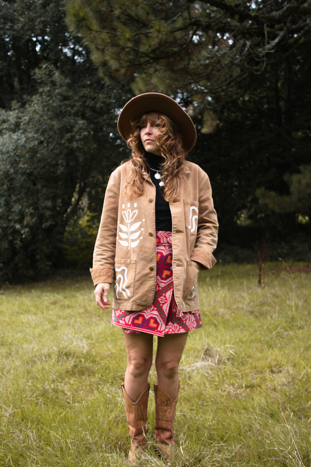 A woman stands in a grassy outdoor setting wearing a tan bohemian-style jacket with white floral embroidery, a wide-brimmed brown hat, a black turtleneck, a red and pink patterned skirt, and brown cowboy boots.