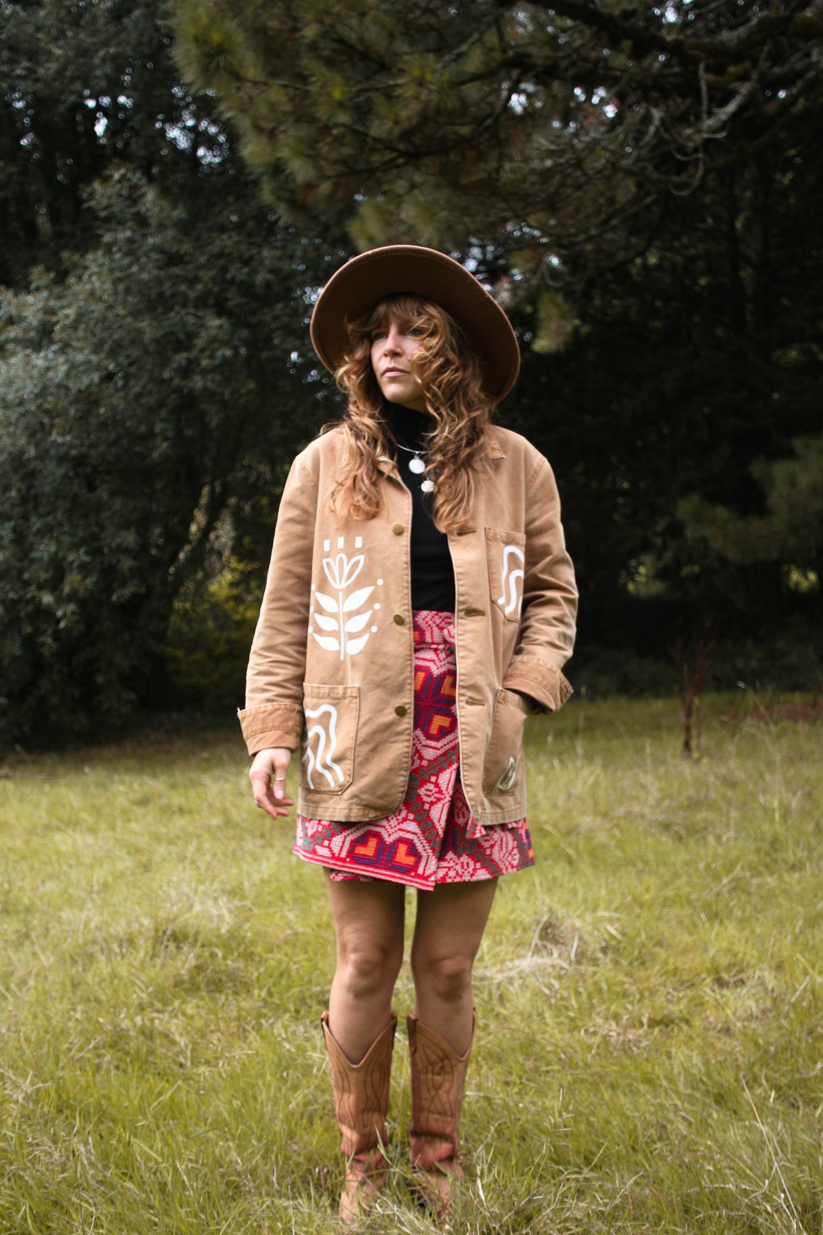 A woman stands in a grassy outdoor setting wearing a tan bohemian-style jacket with white floral embroidery, a wide-brimmed brown hat, a black turtleneck, a red and pink patterned skirt, and brown cowboy boots.