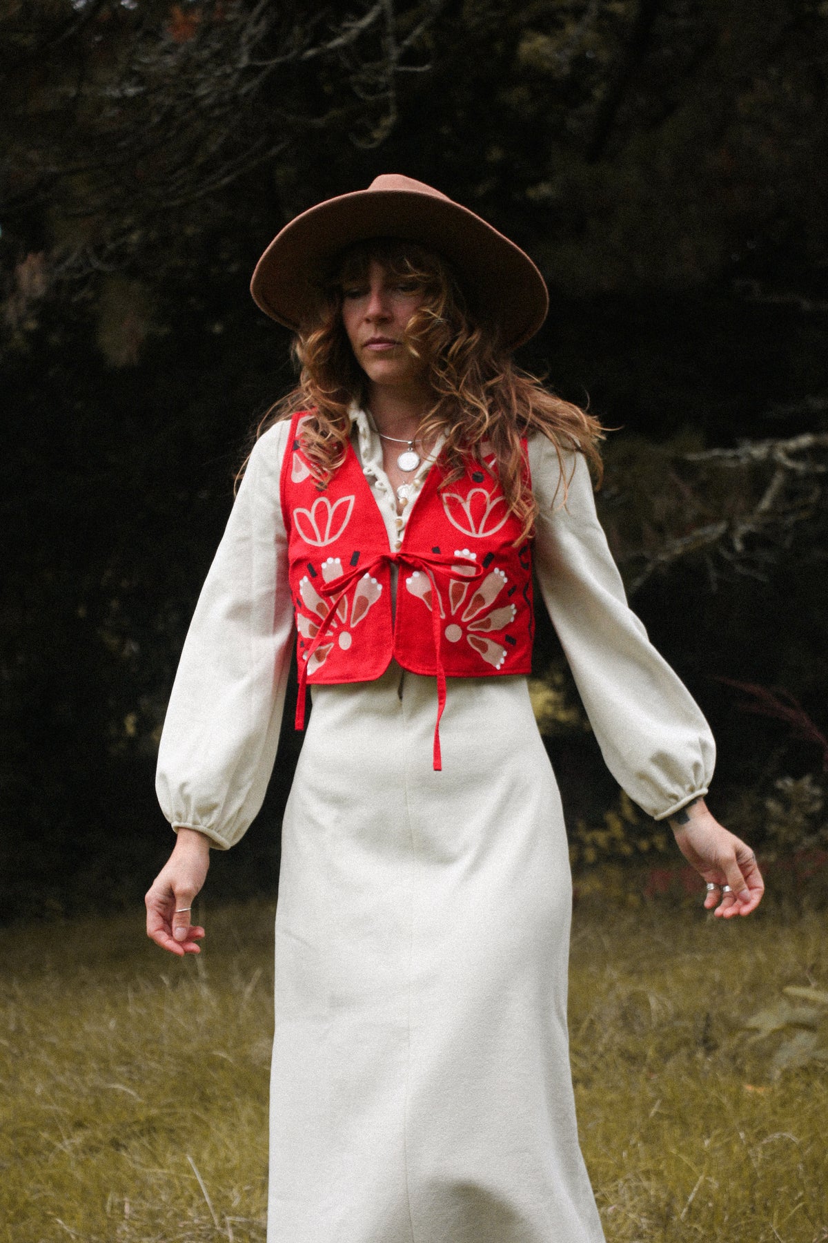 A woman wearing a wide-brimmed brown hat, a cream-colored long-sleeve blouse, a cream maxi skirt, and a red embroidered vest with white floral patterns, standing in an outdoor grassy area with dark trees in the background.