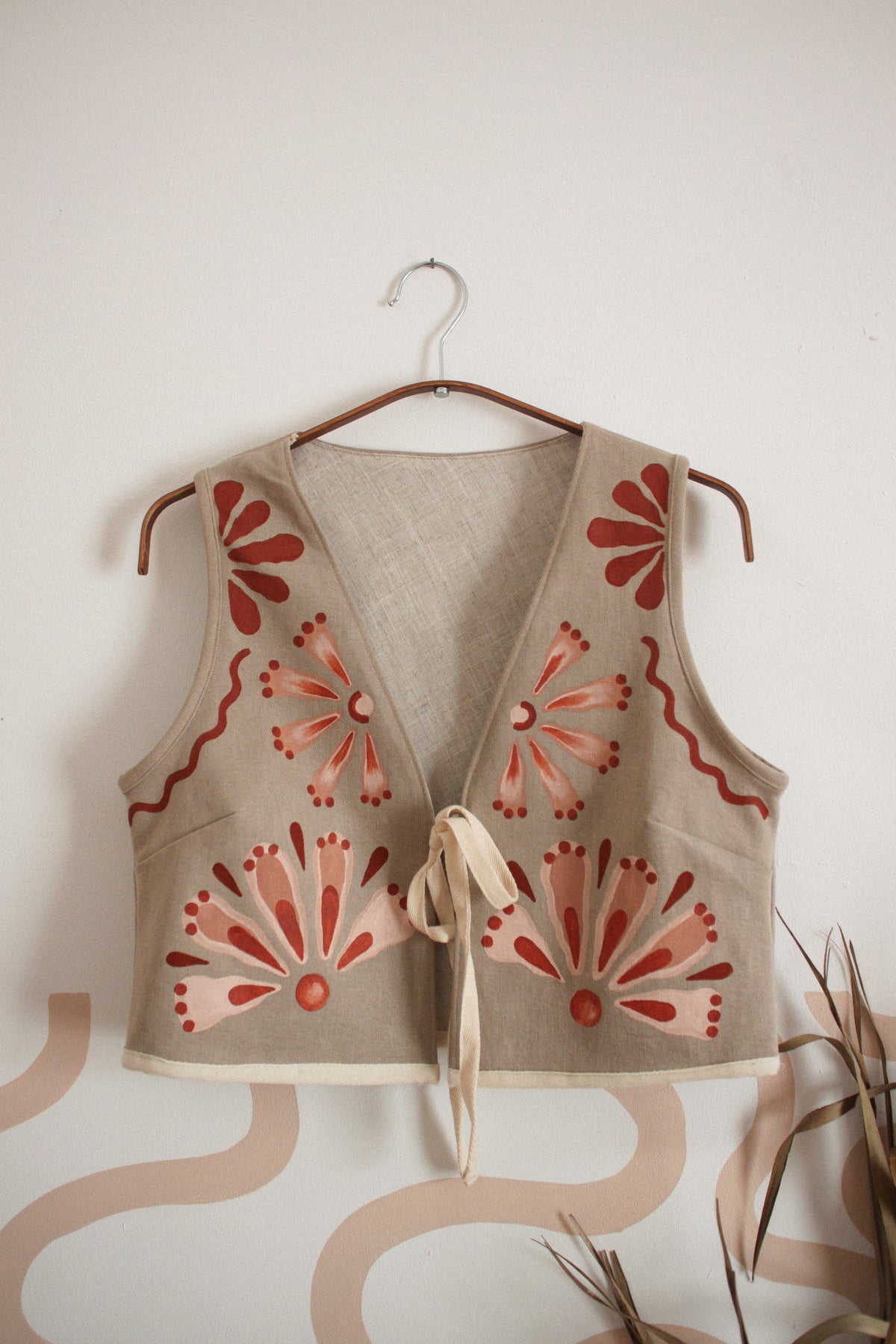 A sleeveless beige vest with red and pink floral patterns, hanging on a wooden hanger against a white wall with decorative wavy wallpaper and dried plants.