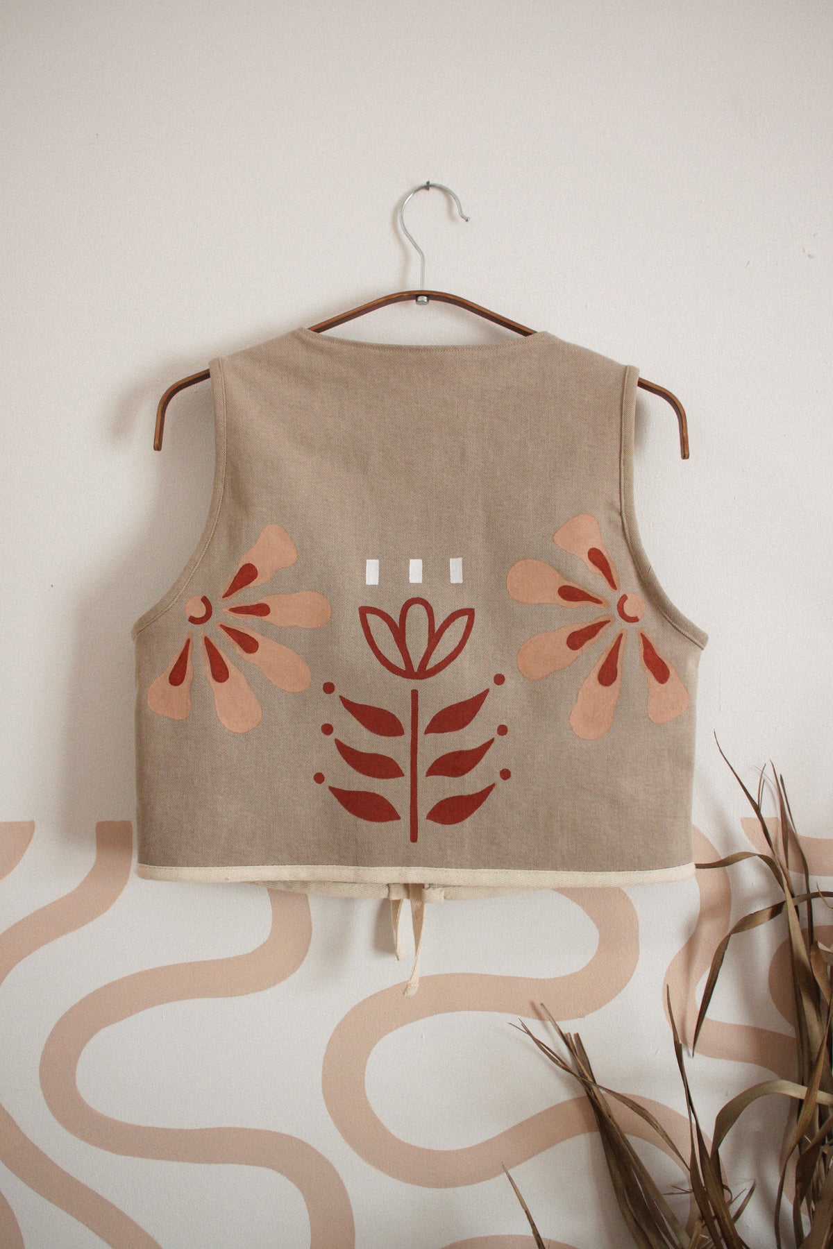 A sleeveless beige linen vest with a hand-painted floral and leaf pattern in terracotta and peach, hanging on a wooden hanger against a white wall with a wavy beige pattern and dried grass in the foreground.