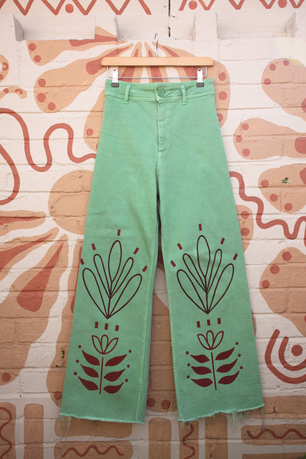 Light green wide-leg cropped jeans with dark brown bohemian floral line-art prints on the front, hanging on a wooden hanger against a patterned brick wall.