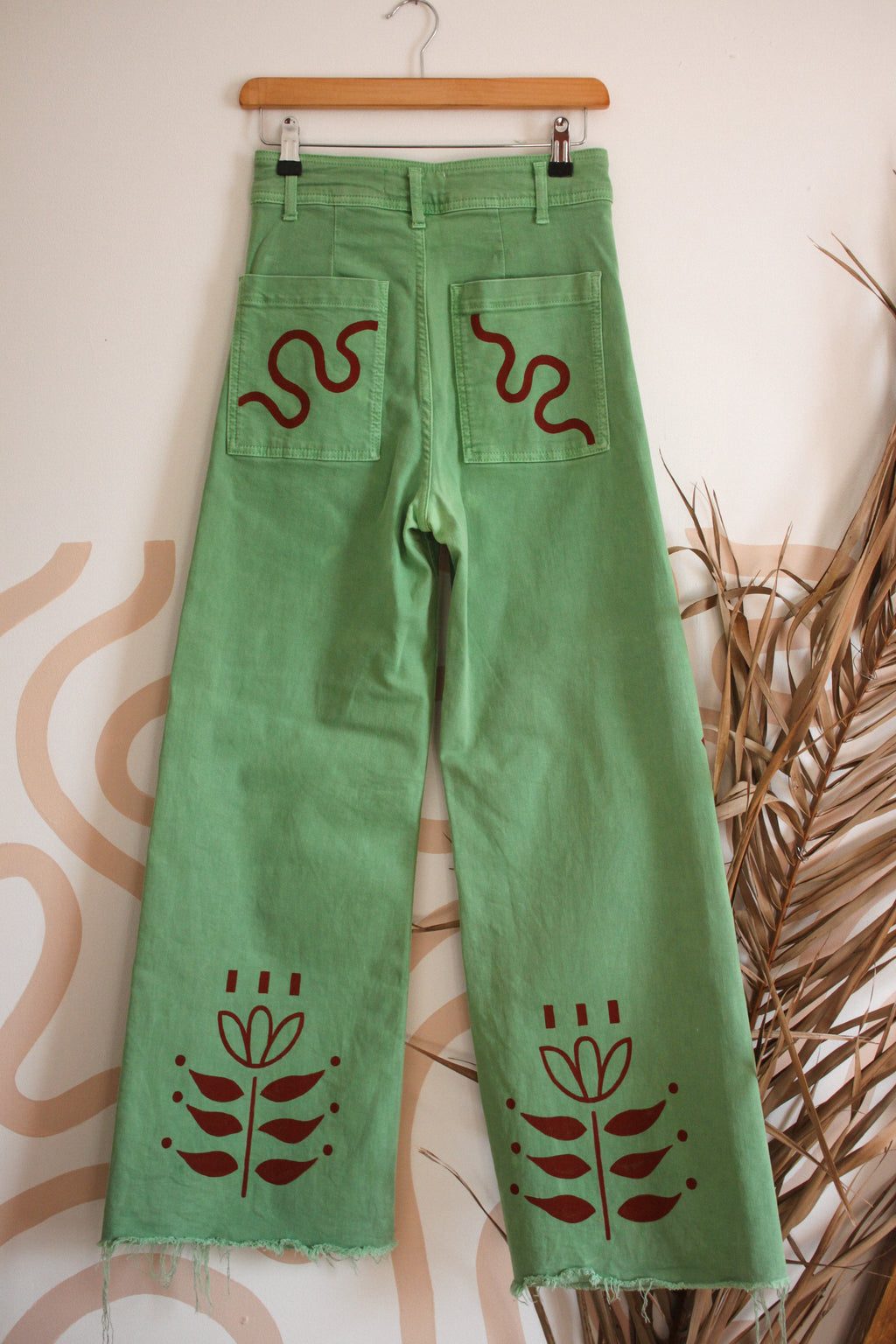 Green wide-leg denim jeans hanging on a wooden hanger against a patterned wall, featuring dark red embroidered snake and floral motifs on the back pockets and lower legs.