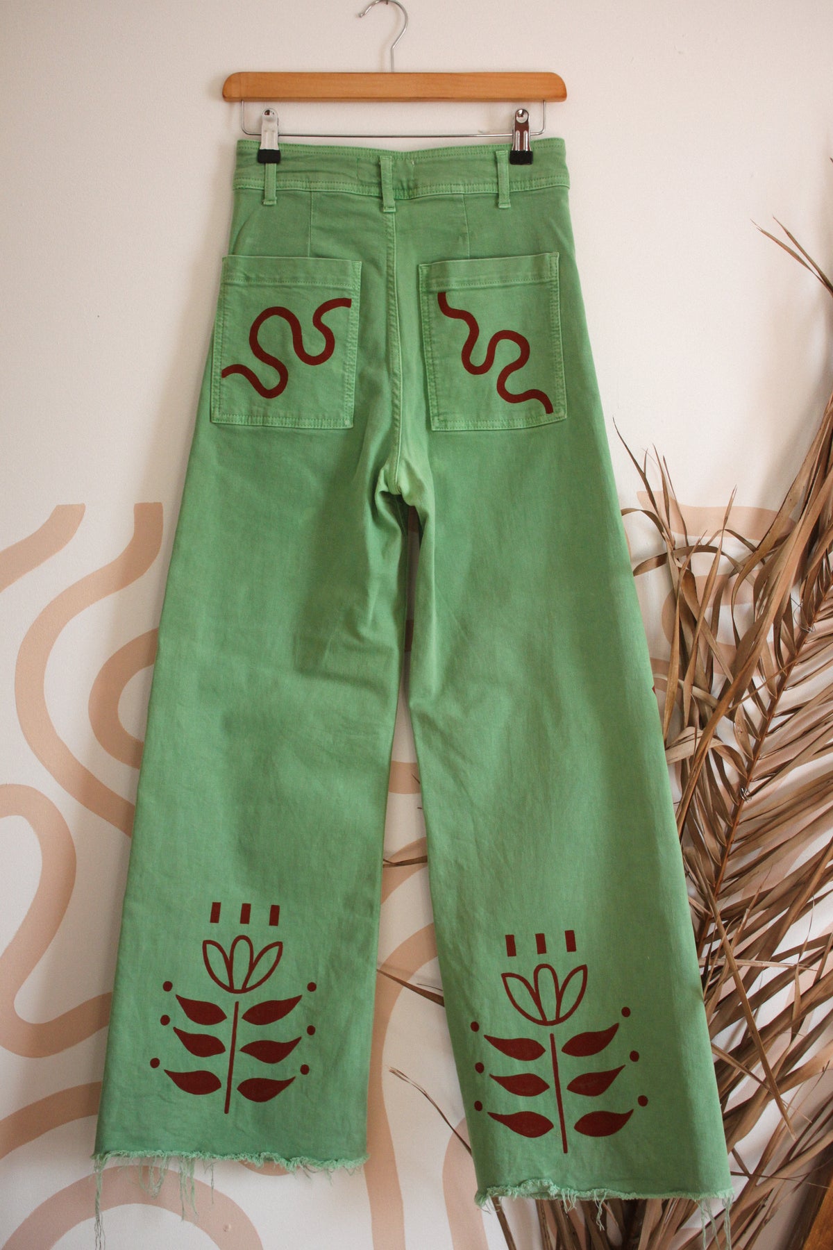Green wide-leg denim jeans hanging on a wooden hanger against a patterned wall, featuring dark red embroidered snake and floral motifs on the back pockets and lower legs.