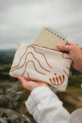 A person outdoors holds a cream canvas pouch with brown minimalist line art and a spiral notebook tucked into the top. The background shows a blurred natural landscape under an overcast sky.