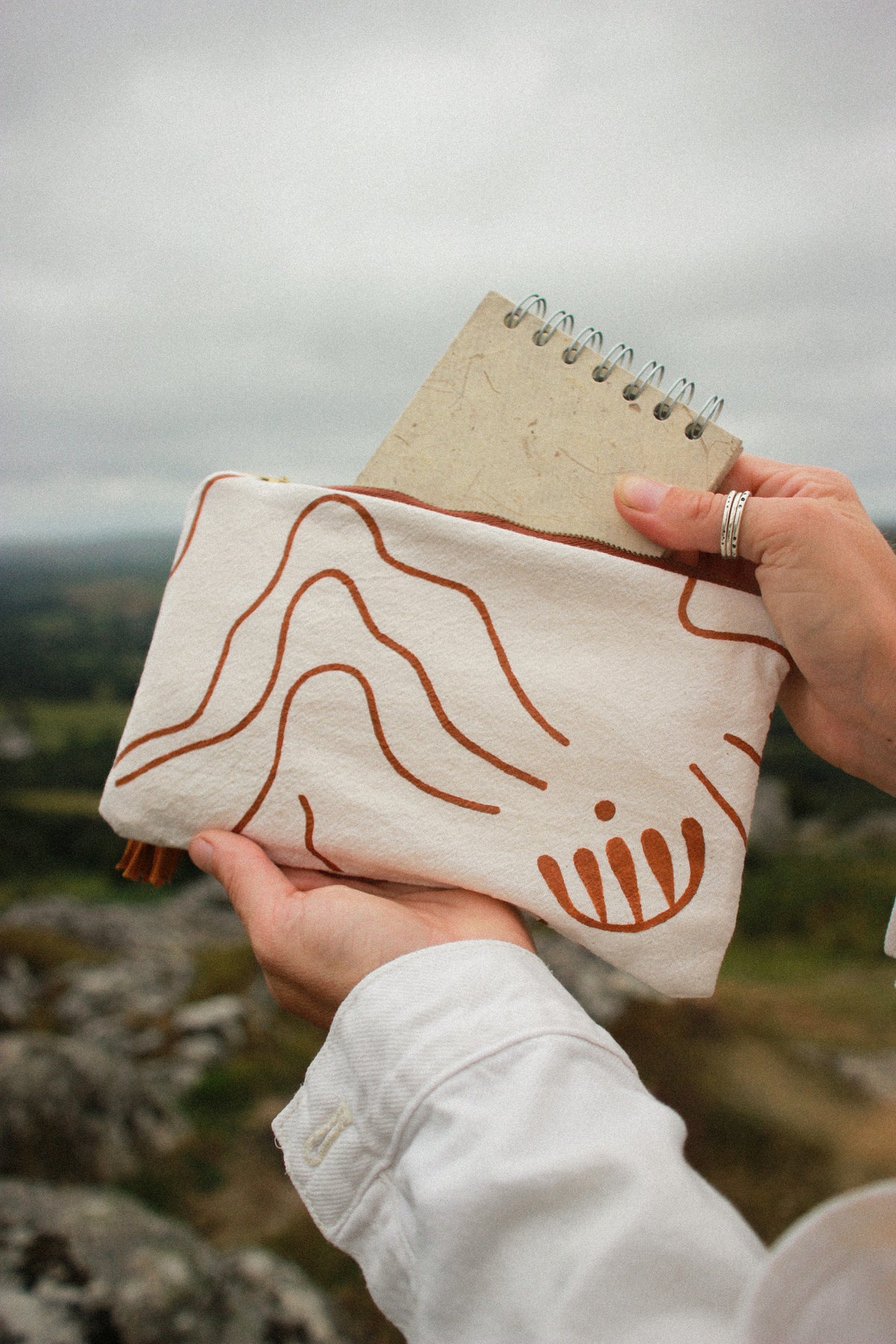 A person outdoors holds a cream canvas pouch with brown minimalist line art and a spiral notebook tucked into the top. The background shows a blurred natural landscape under an overcast sky.