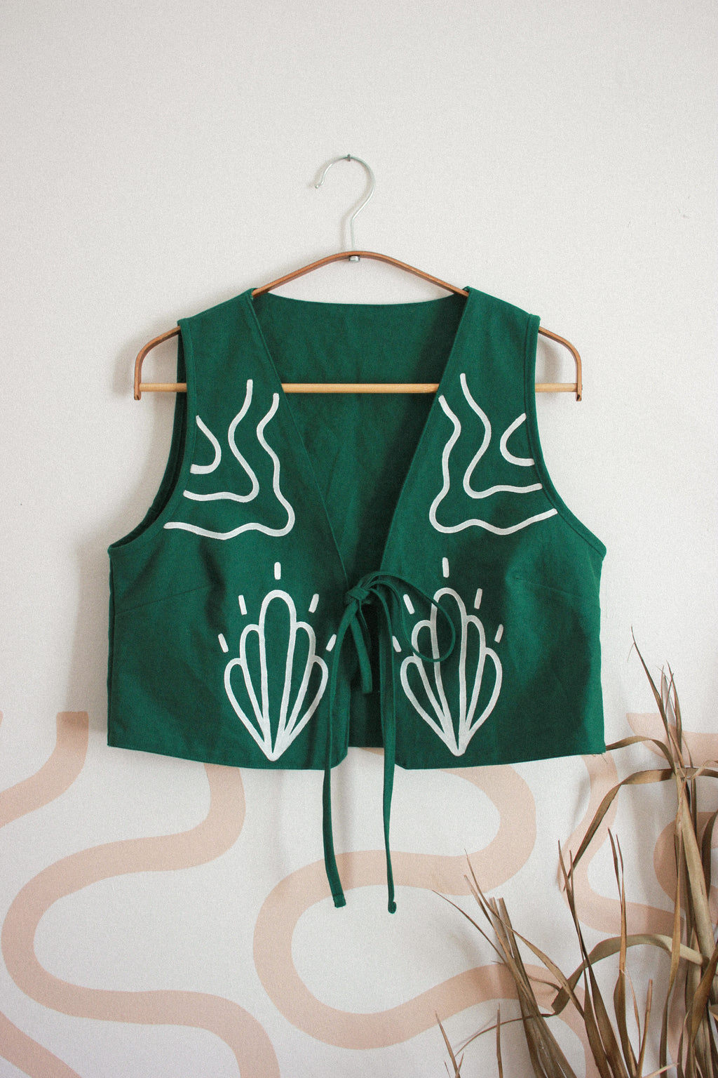 A green sleeveless vest with white abstract line-art prints on the front, hanging on a wooden hanger against a light beige wall with decorative dried grass in the lower right corner.