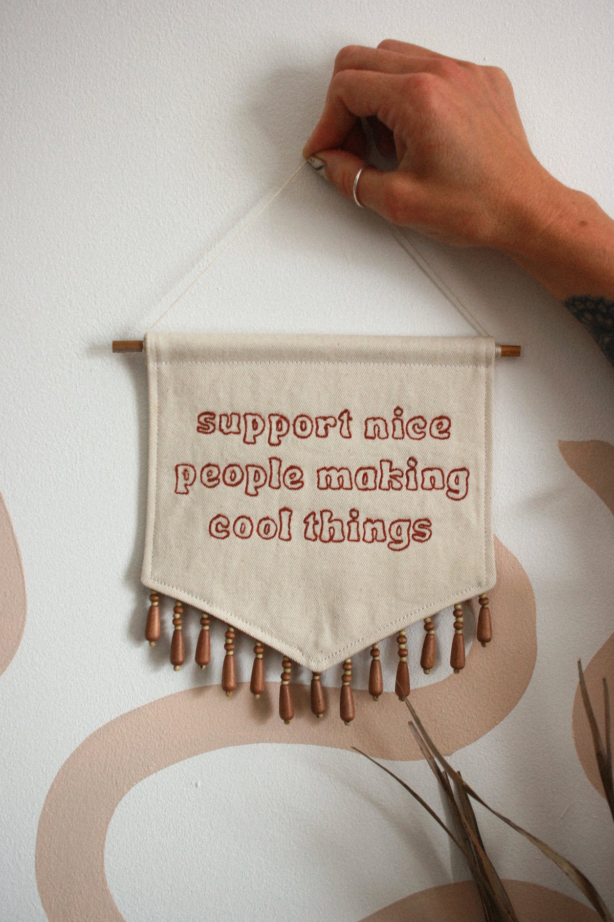 A hand holds a small fabric wall hanging with a wooden rod and fringe. The fabric is off-white with red embroidered text reading 'support nice people making cool things' and brown wooden beads along the bottom edge.