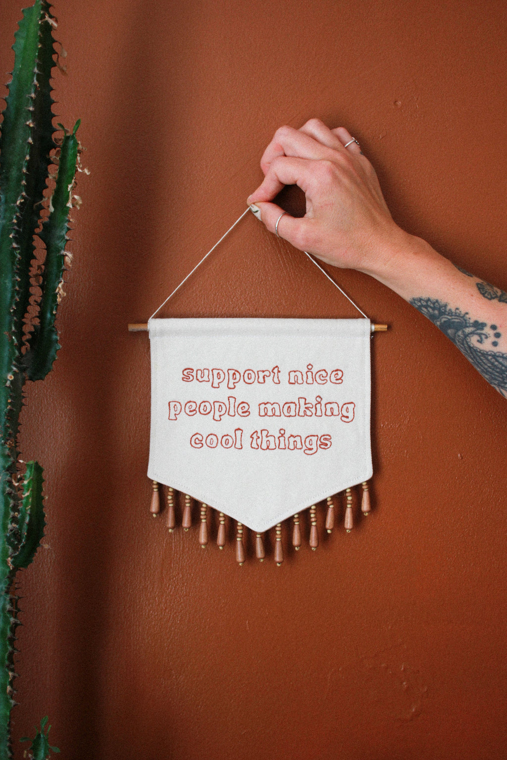 A hand holds a small white fabric wall banner with wooden dowels and fringe, embroidered with the phrase 'support nice people making cool things' in red thread, against a terracotta wall with a cactus on the left.